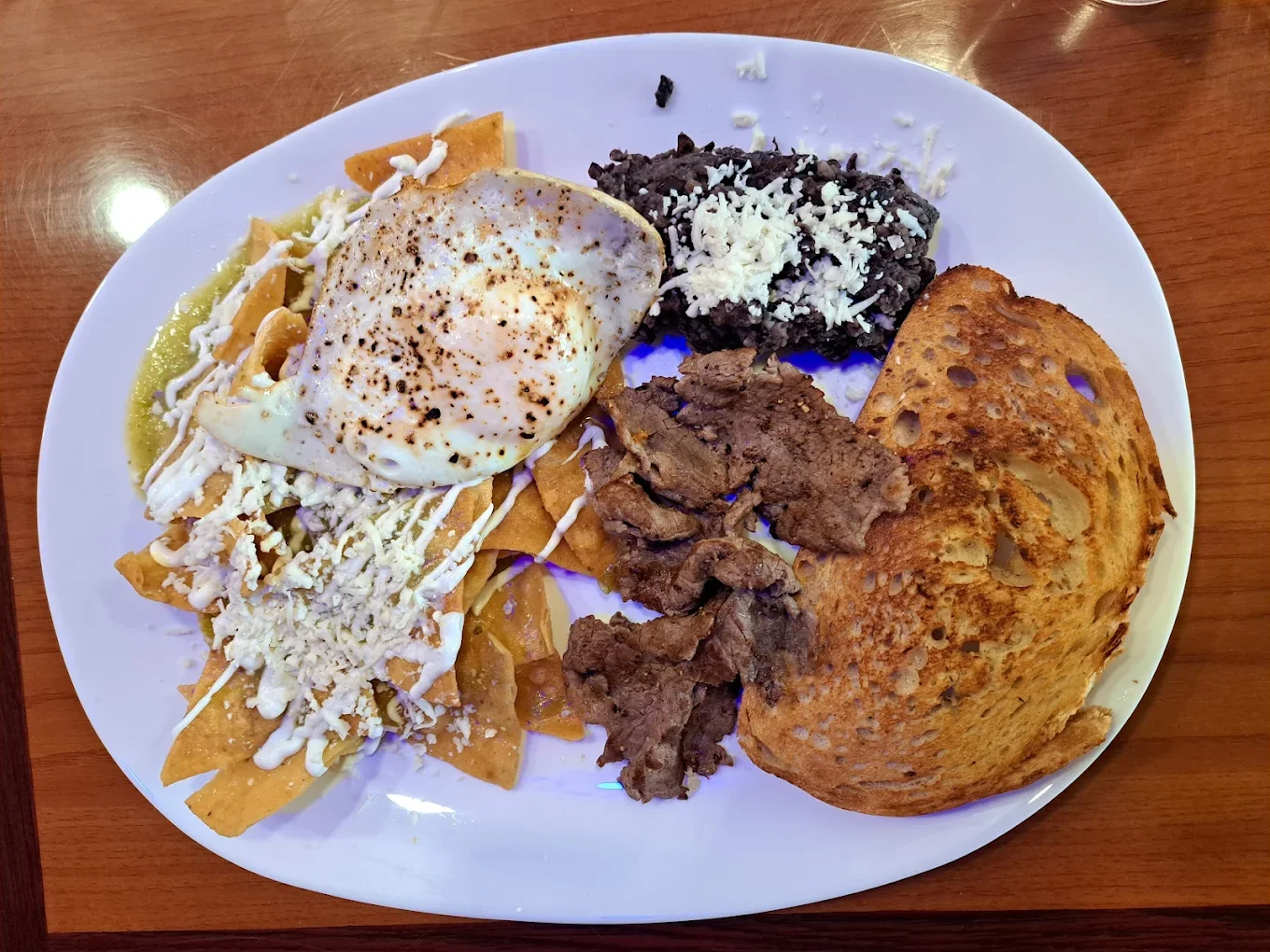 Mexican plate with nachos, a fried egg, carne asada, black beans topped with cheese, and toasted bread on a white oval plate.