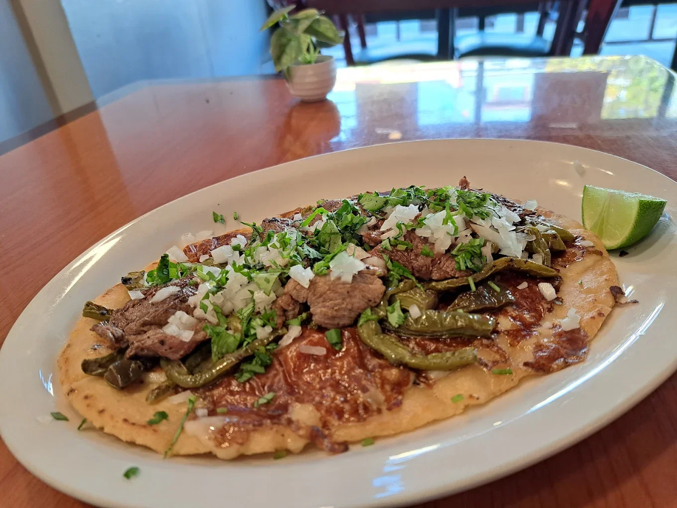 Close-up of a Mexican dish called Tlayuda on a white plate, topped with beef, green chilies, diced onions, cilantro, and a lime wedge, on a restaurant table.