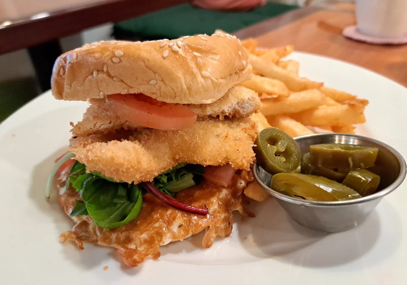 A fried fish sandwich with lettuce, tomato, and tartar sauce on a sesame seed bun served with curly fries and pickled jalapeños