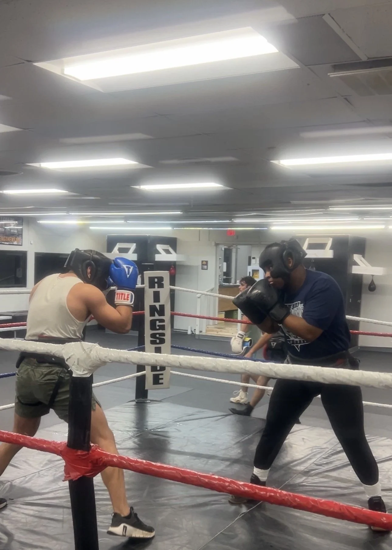 Two boxers with protective headgear and gloves training in a boxing ring, practicing punches and stance, in a gym with bright ceiling lights.