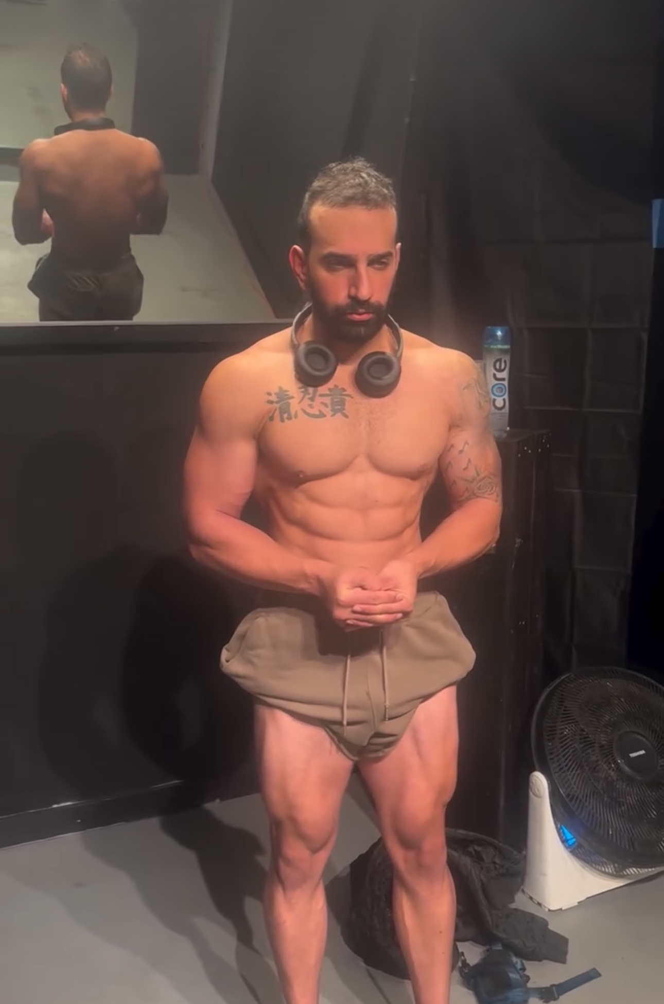 A muscular, shirtless man with headphones around his neck, standing in a room with black walls and a mirror behind him, wearing beige shorts and black shoes, with a water bottle and a fan nearby.