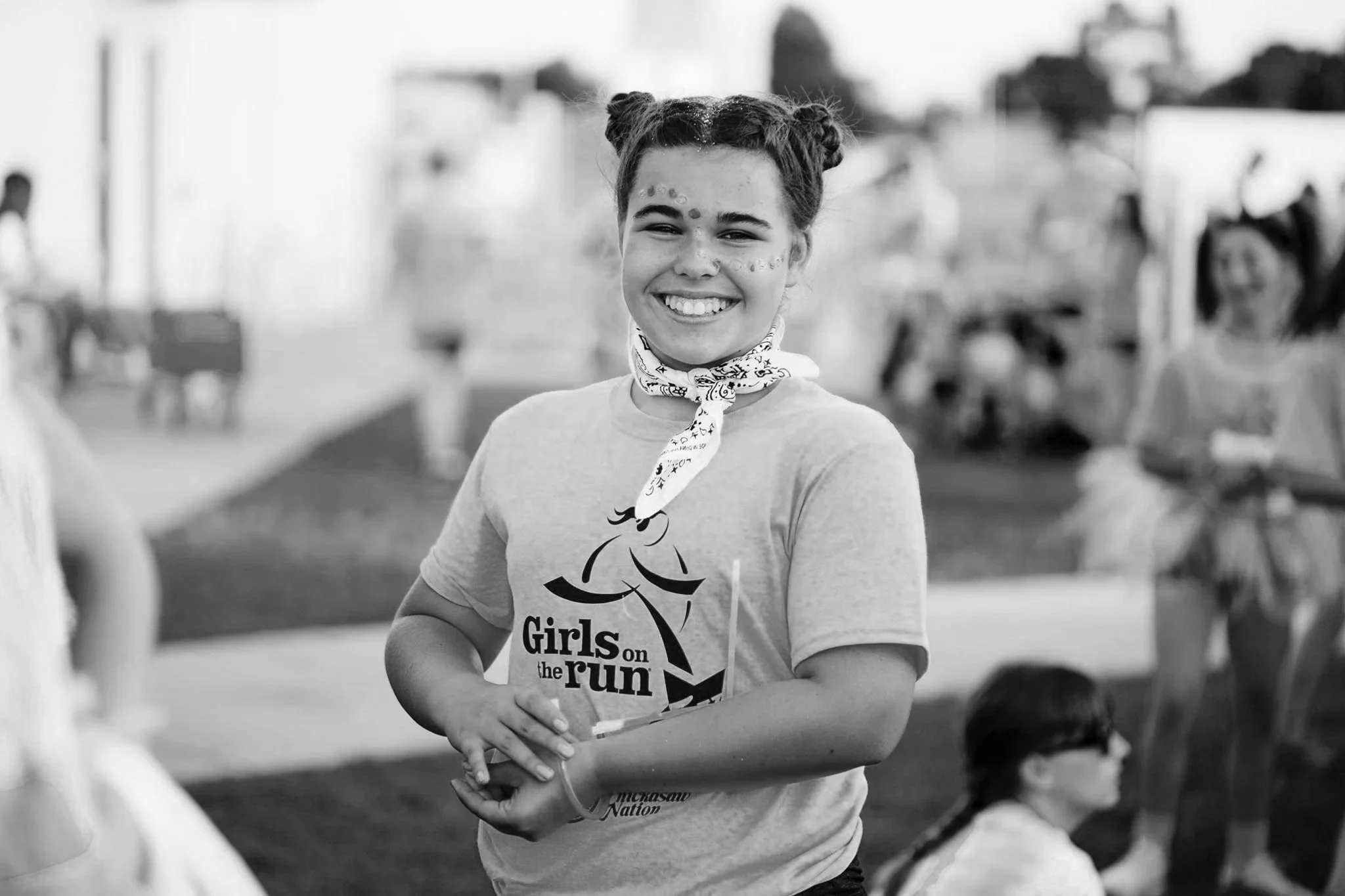 A young girl with braided hair and a bandana around her neck, smiling at the camera, at an outdoor event with blurred people and structures in the background.