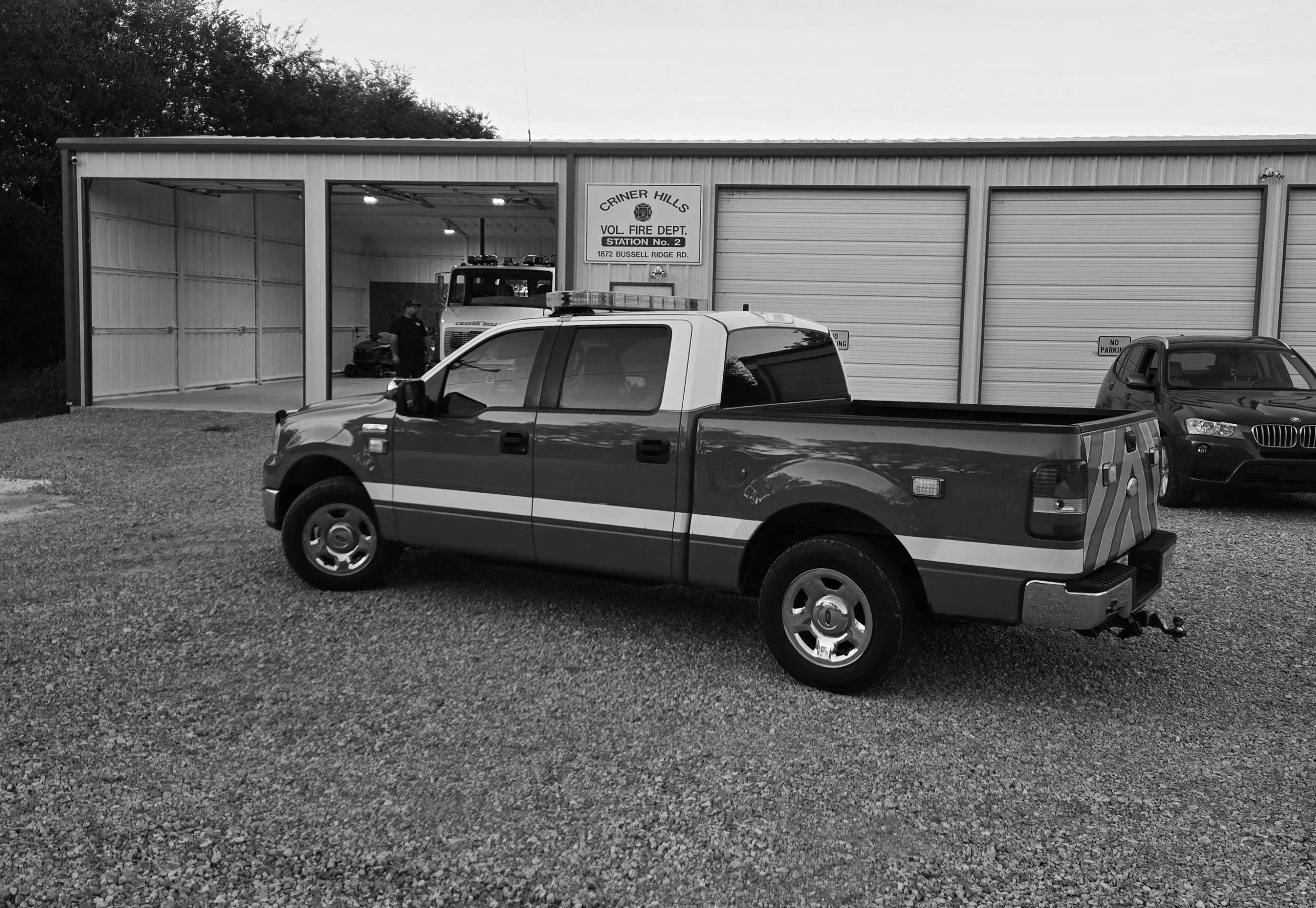 Black and white photo of a fire station garage with a fire truck inside, a pickup truck and a car parked outside, and a sign indicating it is Criner Hills Volunteer Fire Department Station No. 2.
