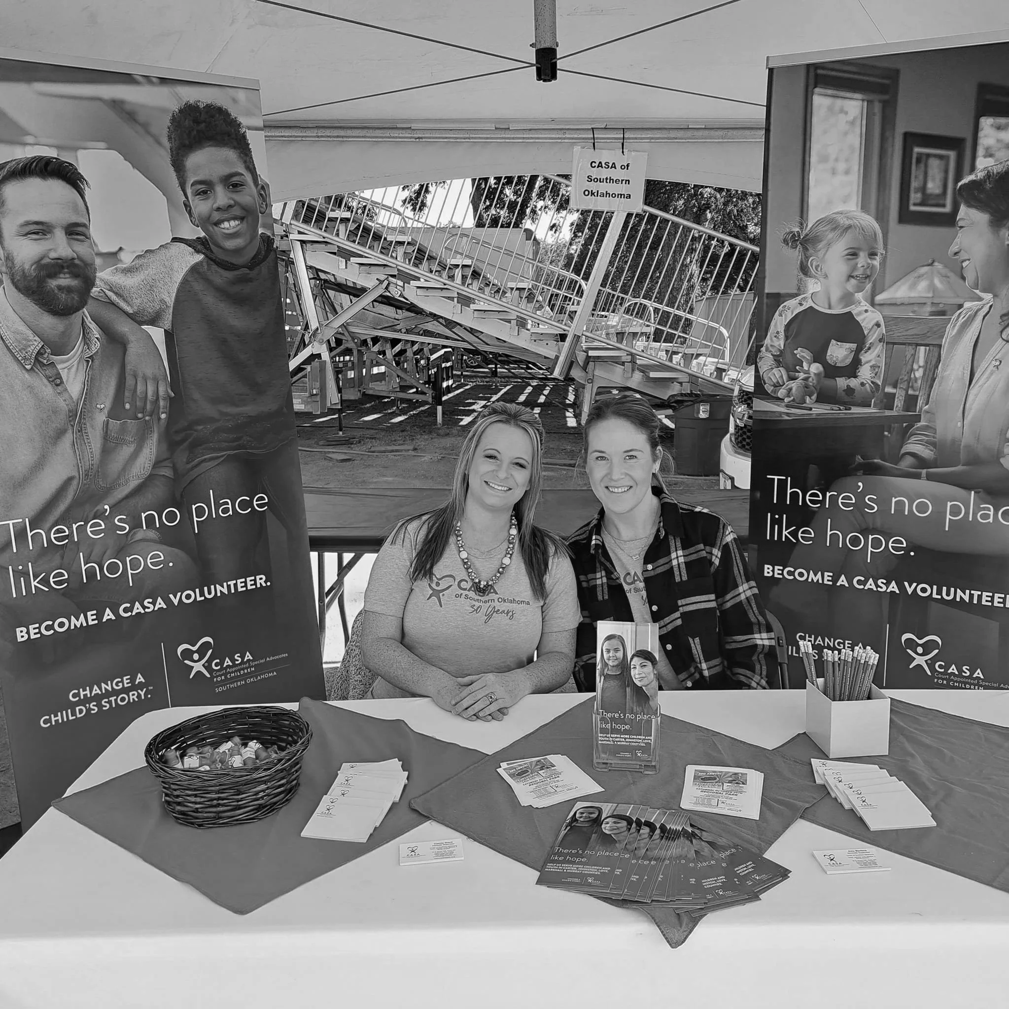 People at an information table promoting CASA volunteer opportunities in Southern Oklahoma. The table has flyers, brochures, a basket, and signs with messages about hope and childhood stories.