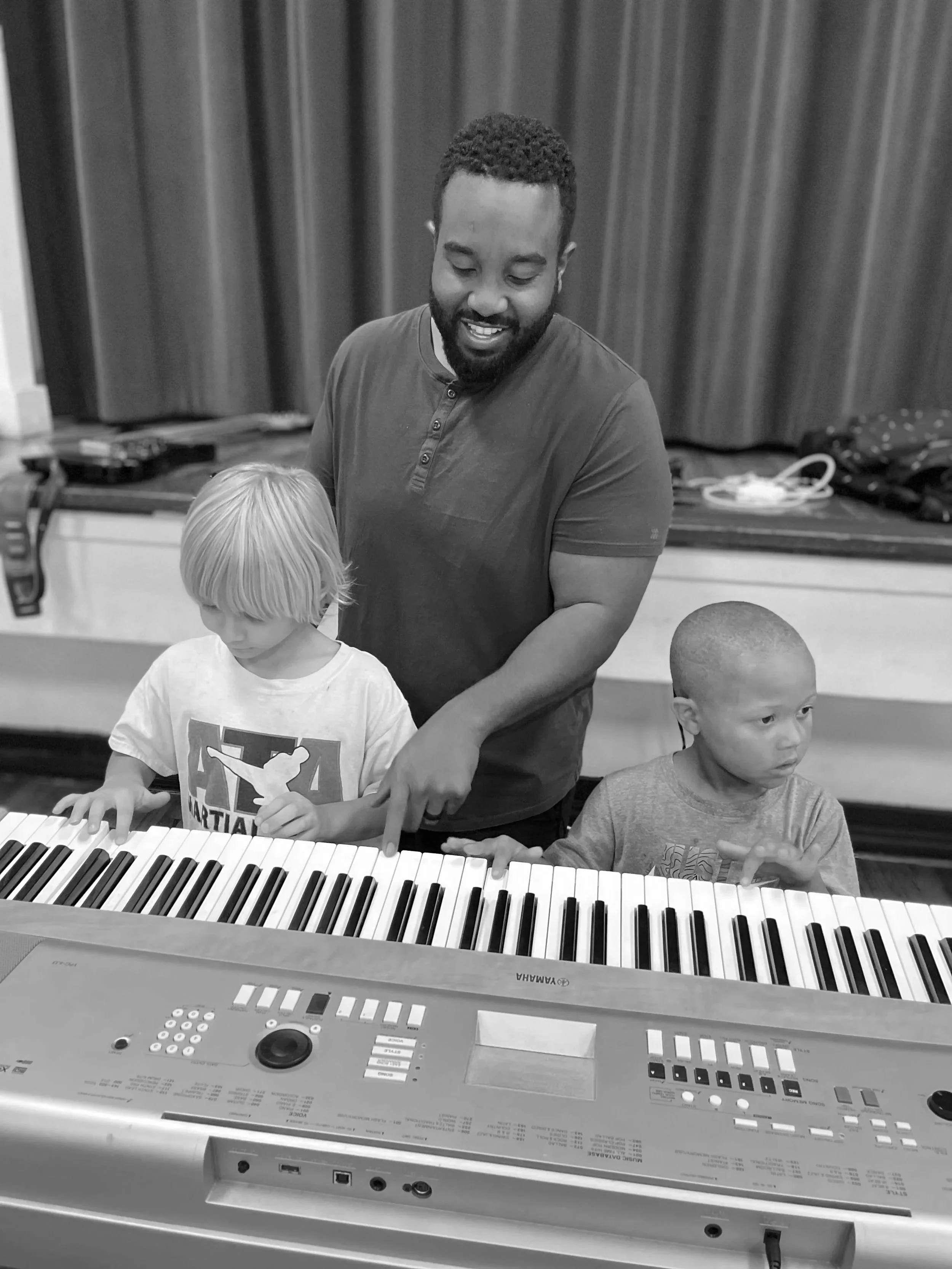 A man with a beard and curly hair smiling while helping two young children play a keyboard in a room with curtains in the background.