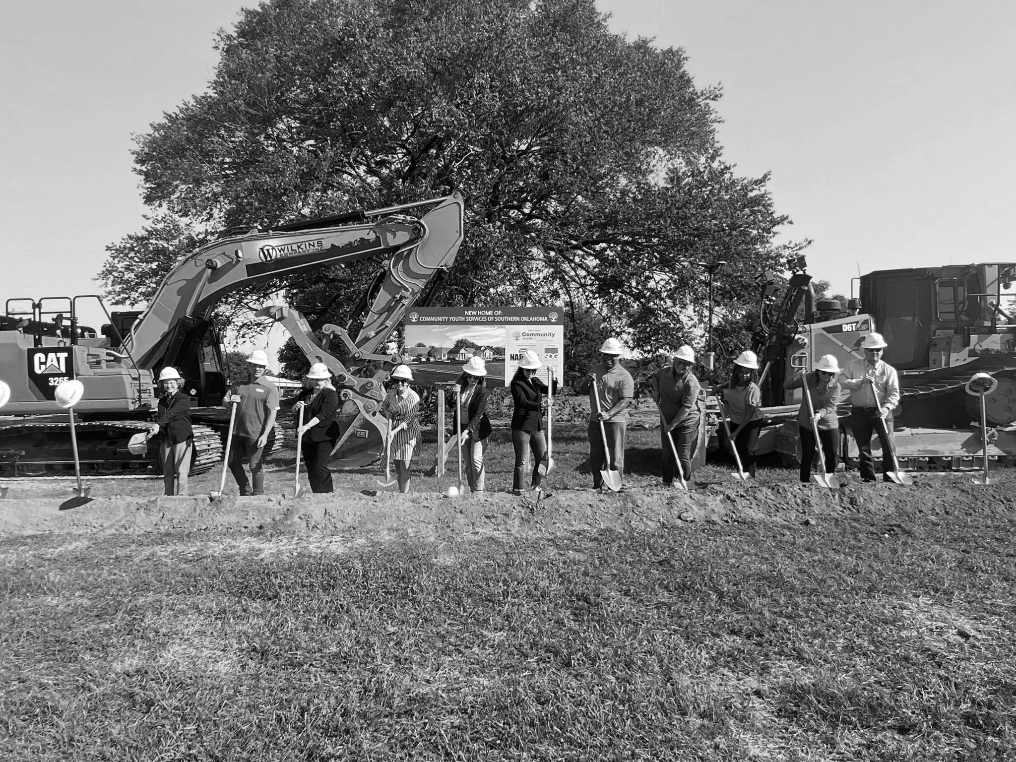 A group of people wearing safety helmets stands in a line in front of construction equipment, including a large excavator, outdoors near a large tree and a sign indicating a new community center in southern Oklahoma.