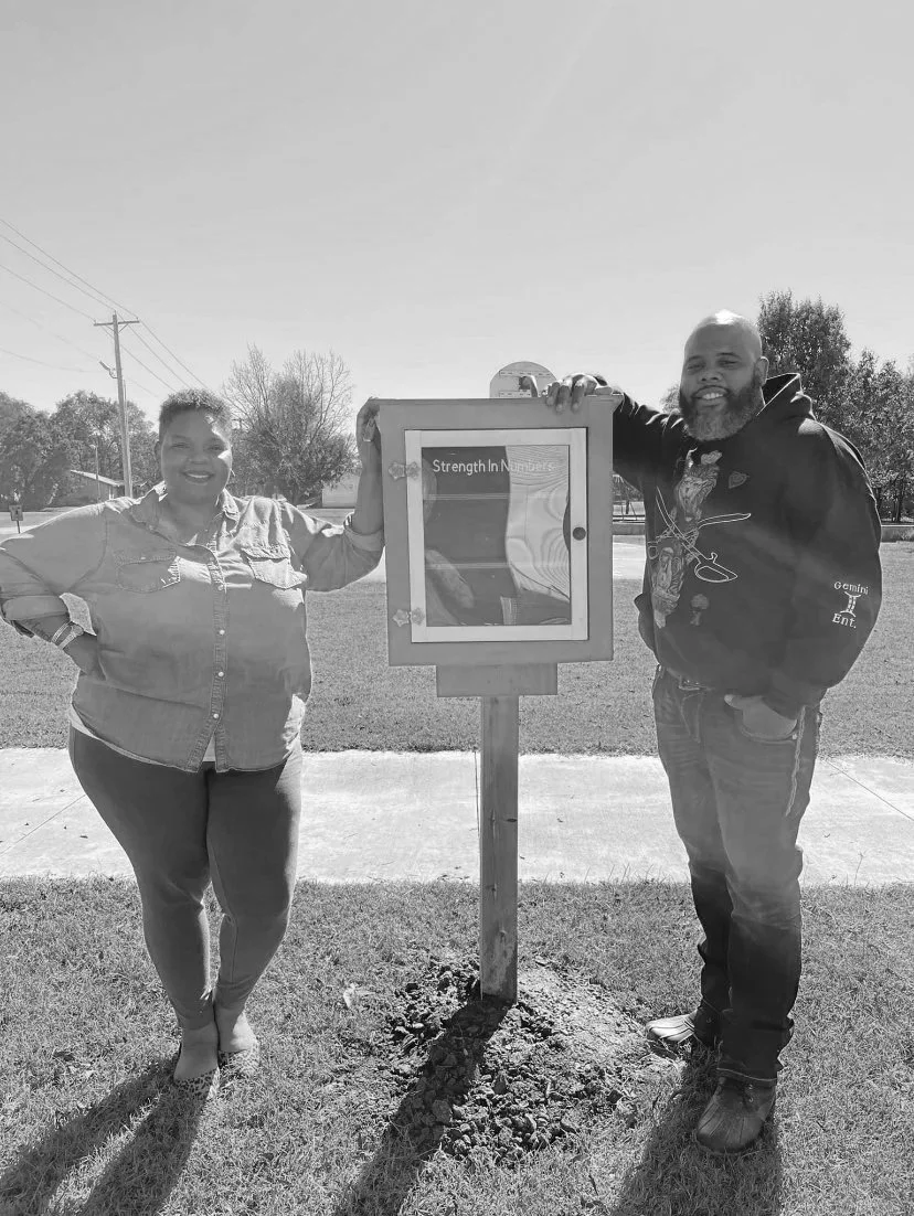 Two people standing outdoors next to a small informational kiosk on a pole. The person on the left is a woman with short hair, wearing a button-up shirt, standing with one hand on her hip. The person on the right is a man with a beard, wearing a hoodie, standing with one hand in his pocket. They are smiling and posing for the photo.
