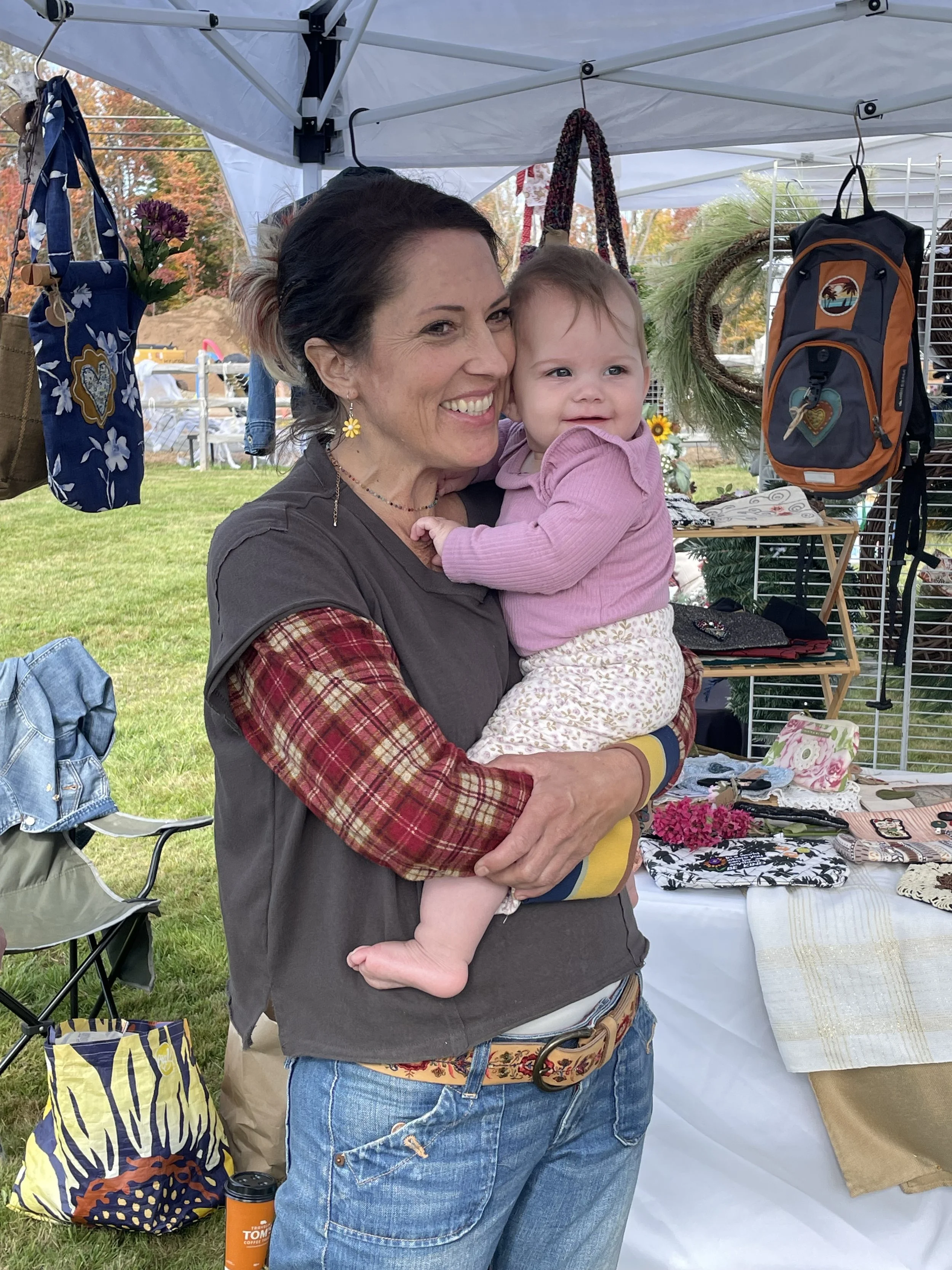 A smiling woman holding a young girl at an outdoor market. The market stalls display bags, wreaths, and other handmade items. The woman is wearing a dark vest over a plaid shirt, jeans, and colorful earrings. The girl is wearing a pink hoodie and patterned pants.