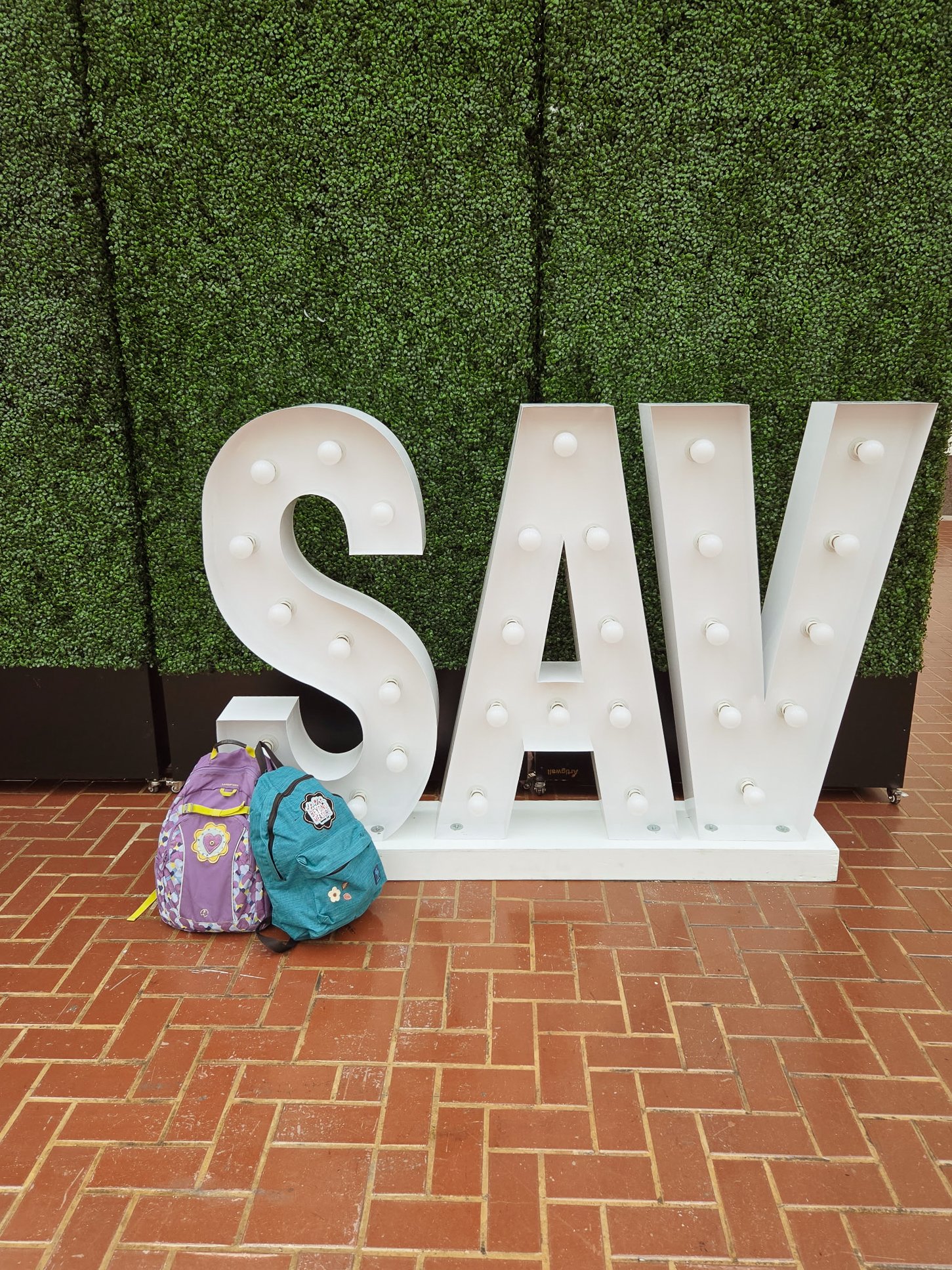 Large white decorative letters spelling 'SAV' with light bulbs on a white base, placed against a green wall made of artificial moss or plants, with two colorful backpacks on a brick floor in front.