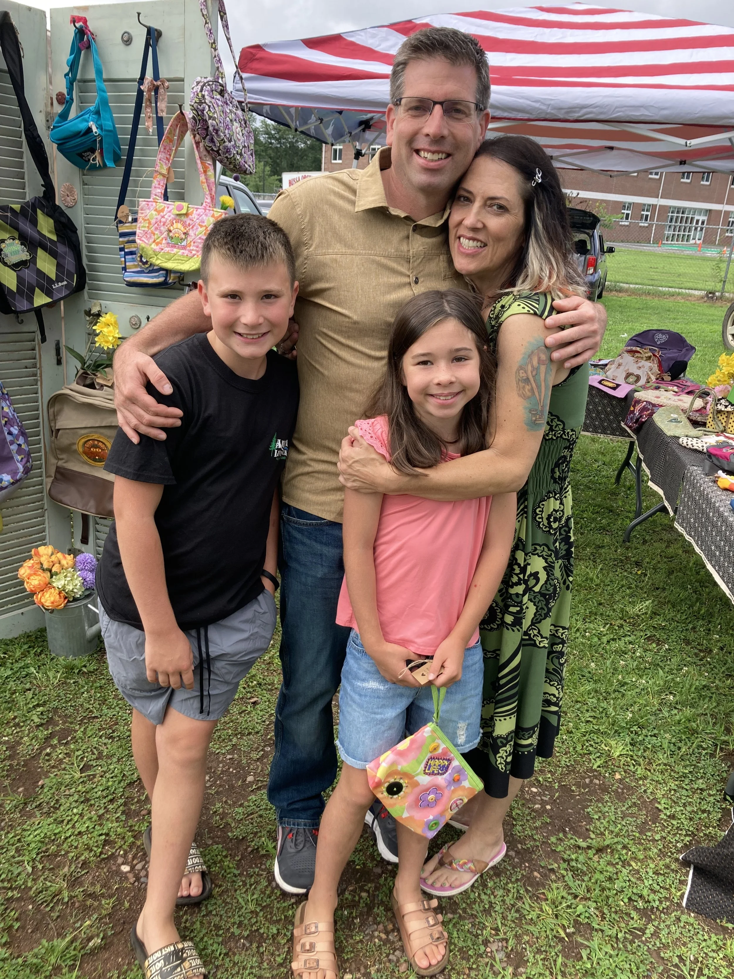 A family of four smiling, standing outdoors at a fair or market under a red and white striped canopy. There are bags hanging on a display and tables with various items behind them. The ground is grassy with patches of dirt.