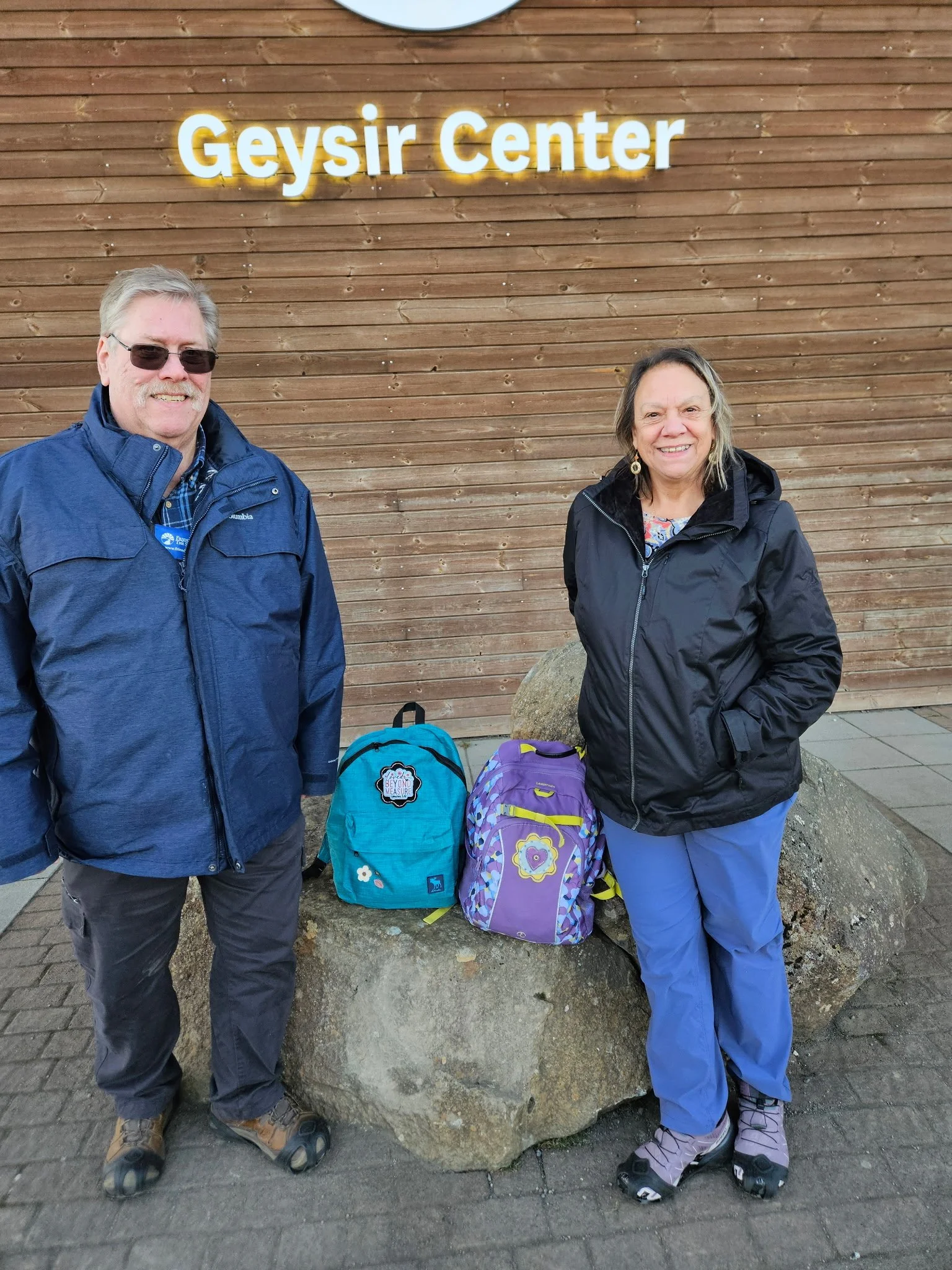 A man and woman standing outdoors in front of a wooden wall with a sign reading 'Geysir Center.' They are smiling, wearing outdoor clothing, and have backpacks with them. The man is on the left wearing sunglasses, a blue jacket, and gray pants. The woman is on the right wearing a black jacket, blue pants, and hiking boots.