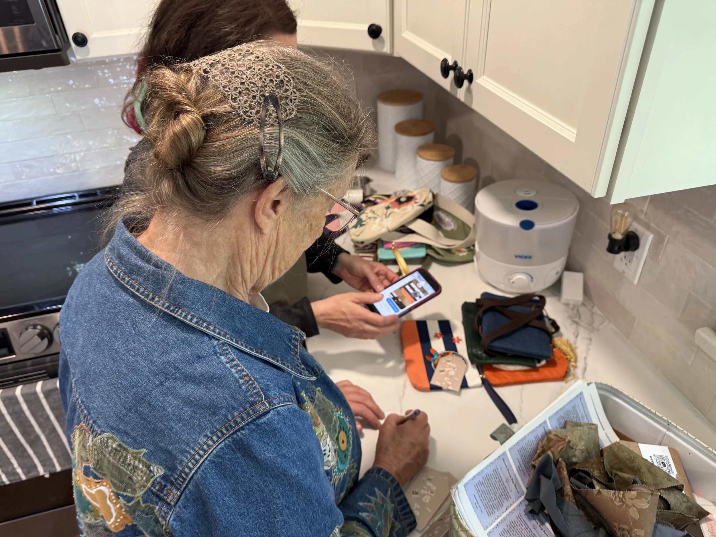 An elderly woman with gray hair and glasses, wearing a denim jacket, is looking at her phone while standing in a kitchen. There are various items on the kitchen counter, including a brochure, a backpack, and a display with informational materials.