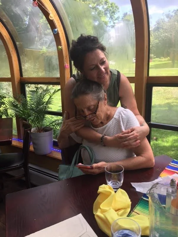 A woman hugging an elderly woman at a table in a sunny, plant-filled space, with the elderly woman looking at her phone.