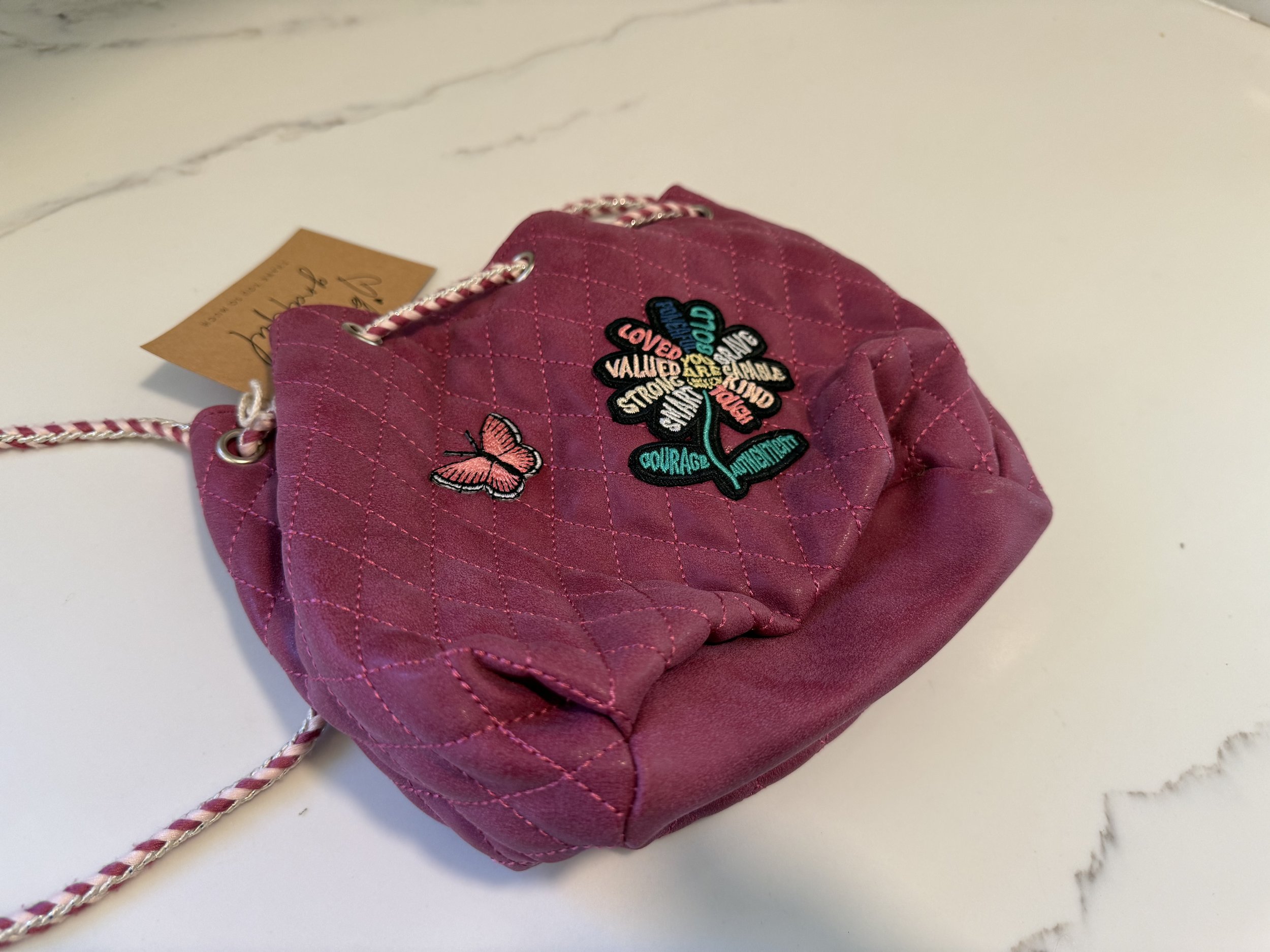 Pink quilted handbag with embroidered flower design and words like 'Loved', 'Valued', 'Courage', and butterflies, on a white surface.