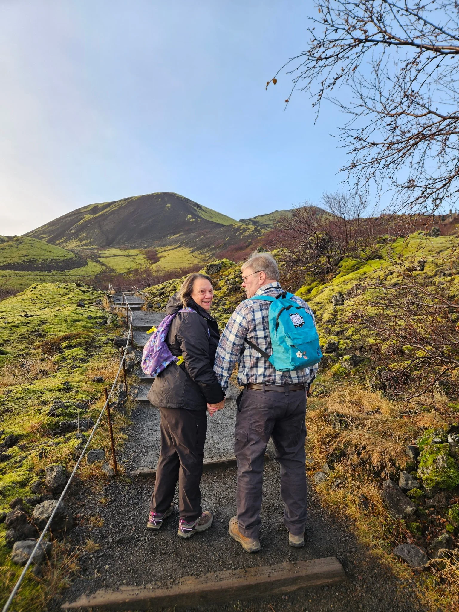 Two hikers, a woman and a man, holding hands and smiling at each other on a mountain trail surrounded by green moss and sparse trees, with a mountain and blue sky in the background.