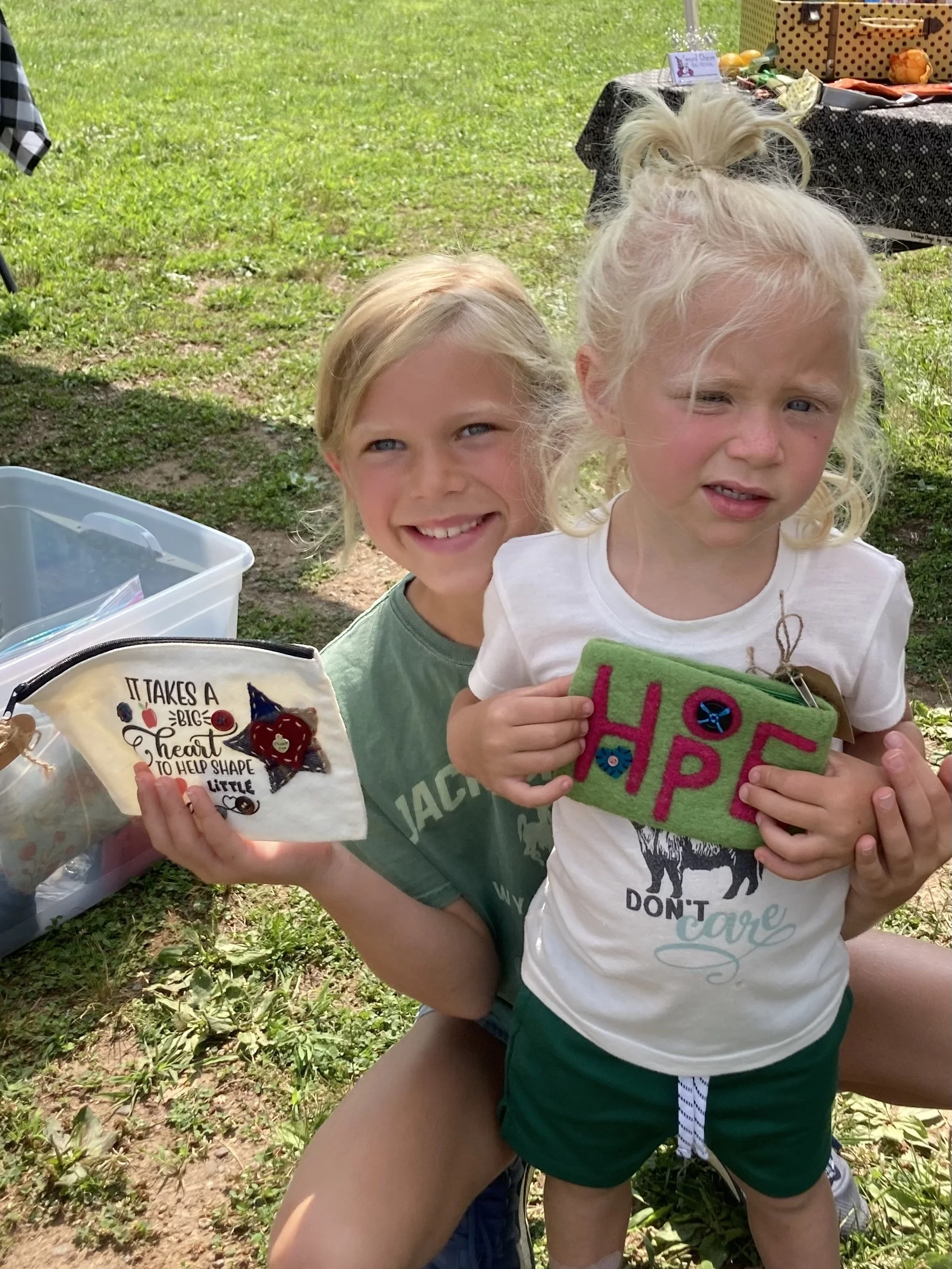 Two young children standing outdoors on grass, holding colorful handmade felt items with words and decorations. One girl has blonde hair tied up and is smiling, while the other has braided blonde hair and appears to be frowning. There are tables with various items and a plastic container nearby.