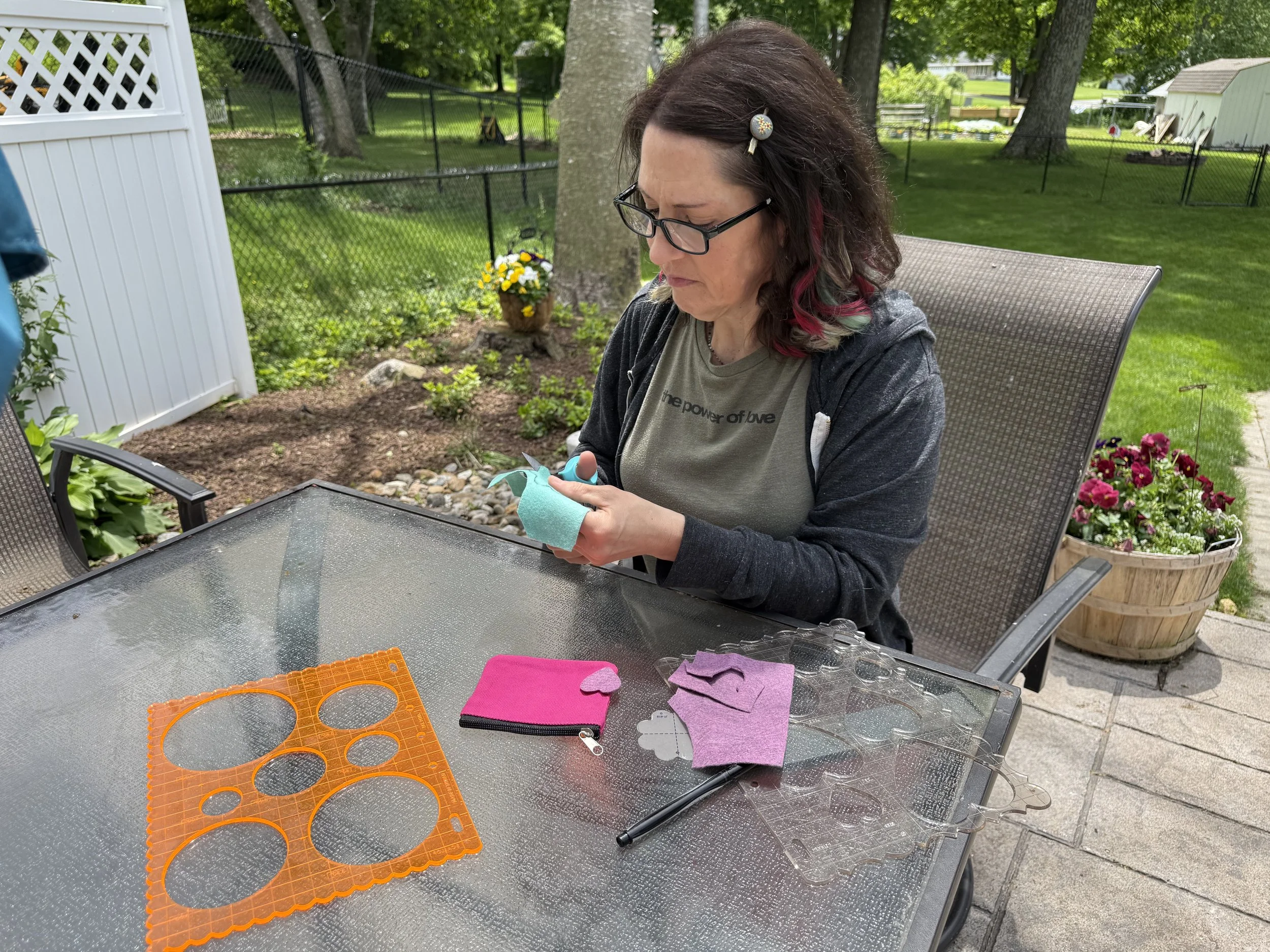 Woman sitting at an outdoor table, cutting felt with scissors, surrounded by craft supplies and colorful flowers in a garden.