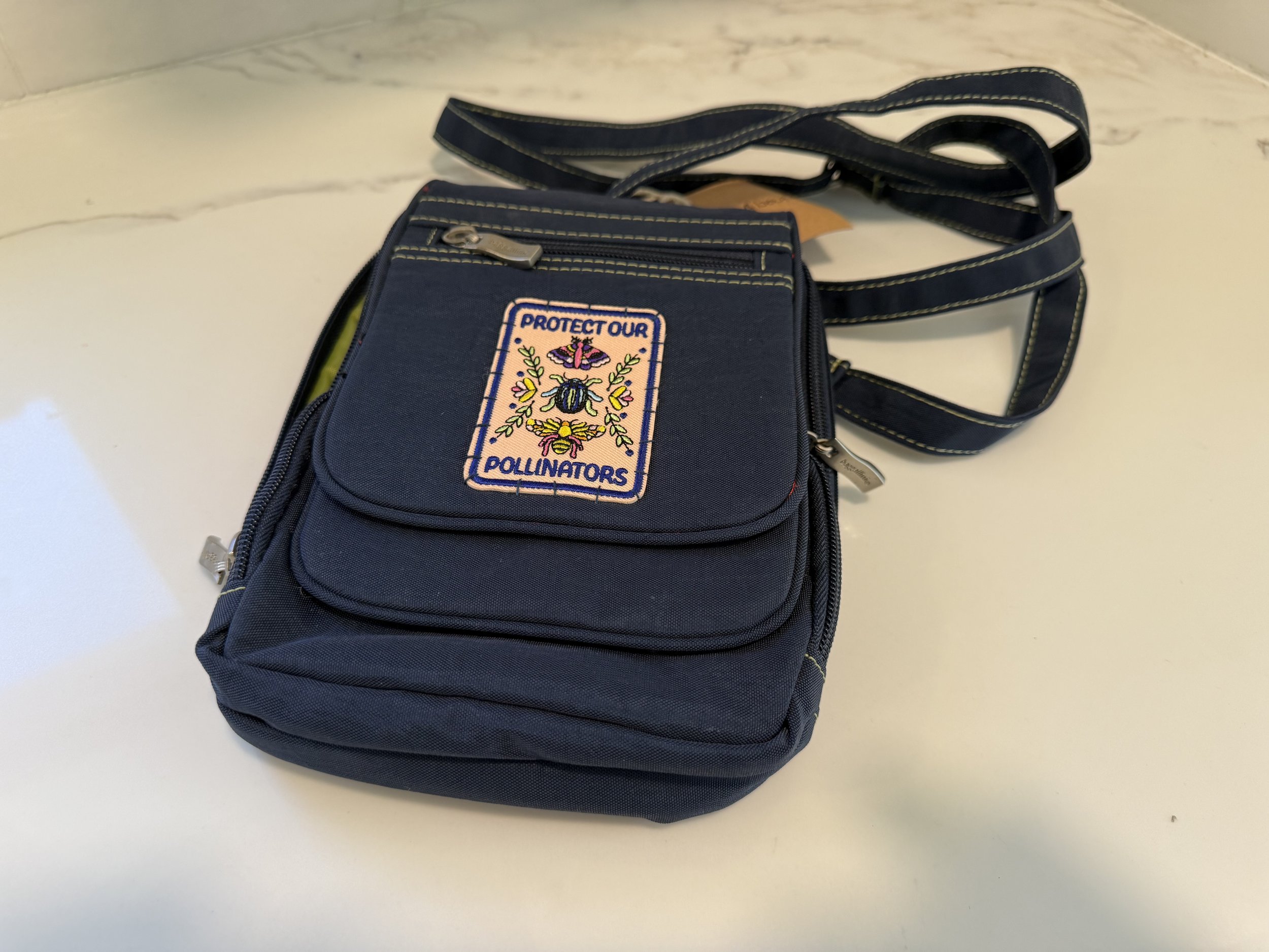 A navy blue school bag with a patch that reads 'Protect Our Pollinators' and an embroidered bee and flowers. The bag has multiple zippered compartments and a strap, placed on a white marble surface.