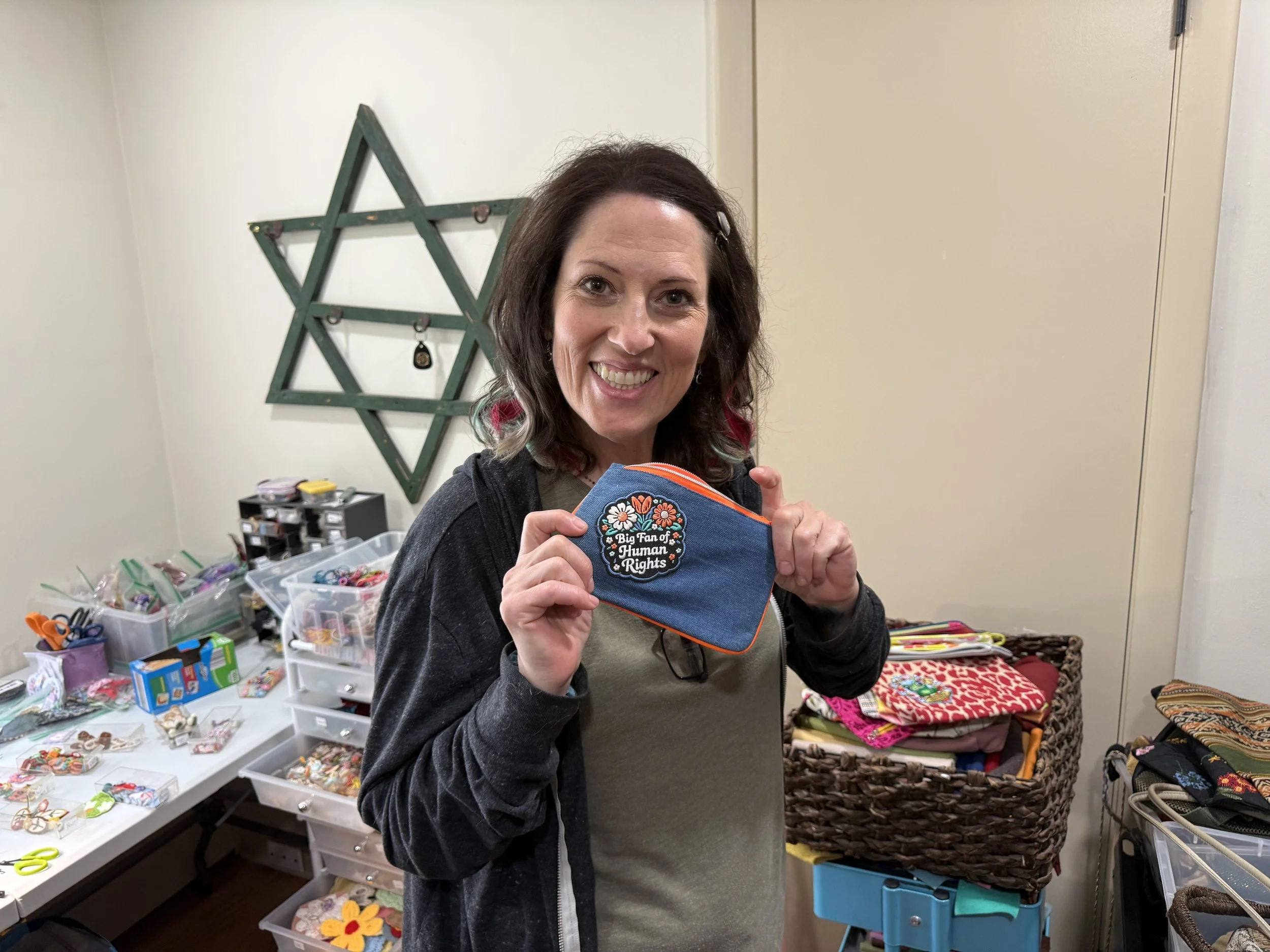 A woman smiling at the camera holding a small fabric pouch with floral embroidery that reads 'Big Fan of Human Rights'. She is in a room with various craft supplies, including baskets of fabric, finished crafts, and containers of small decorative items.