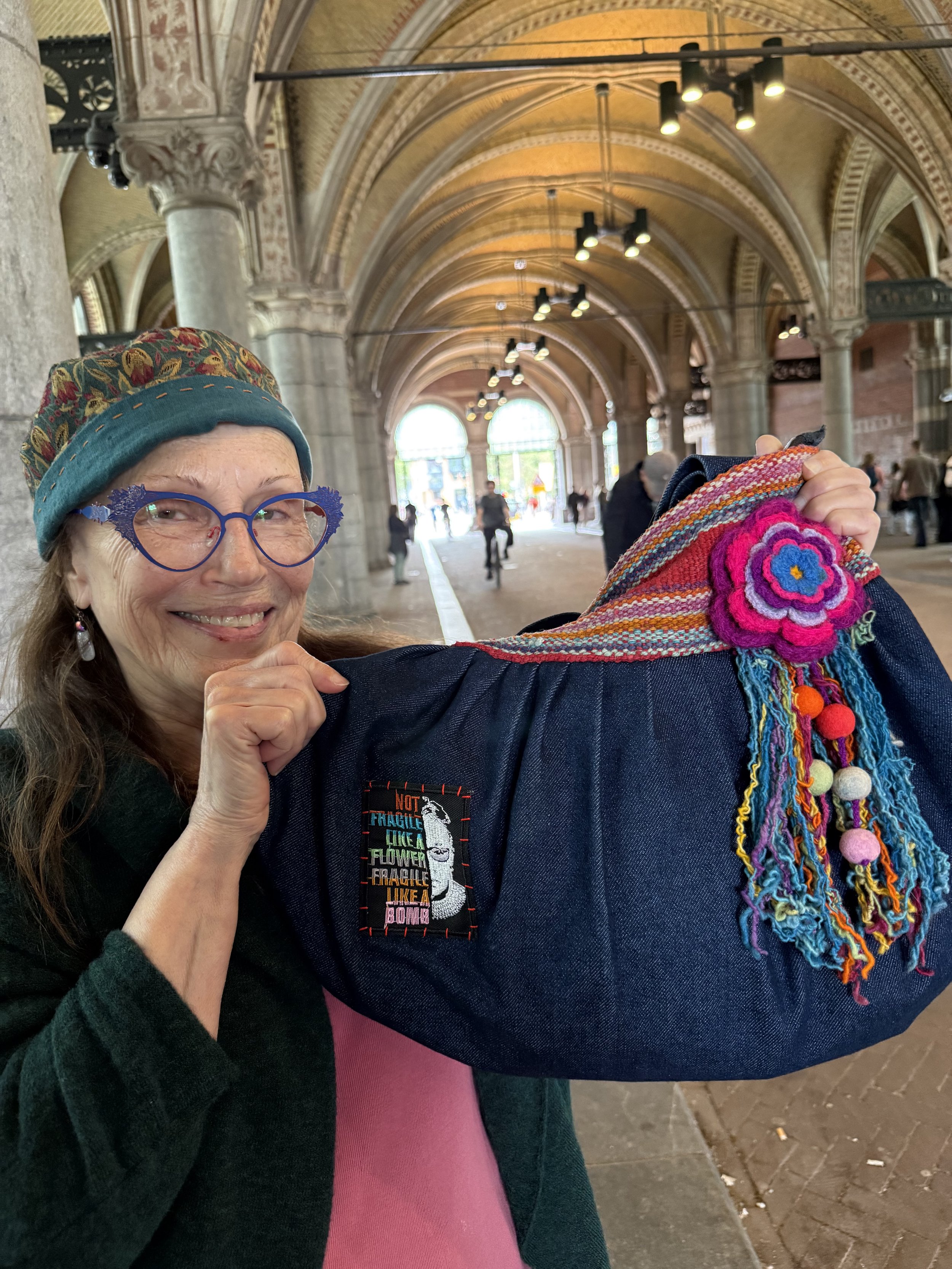 A smiling woman wearing colorful glasses, a patterned hat, and a dark jacket holds a person's arm with a colorful, handmade fabric and felt flower accessory inside a large, ornate building with arched ceilings and people in the background.