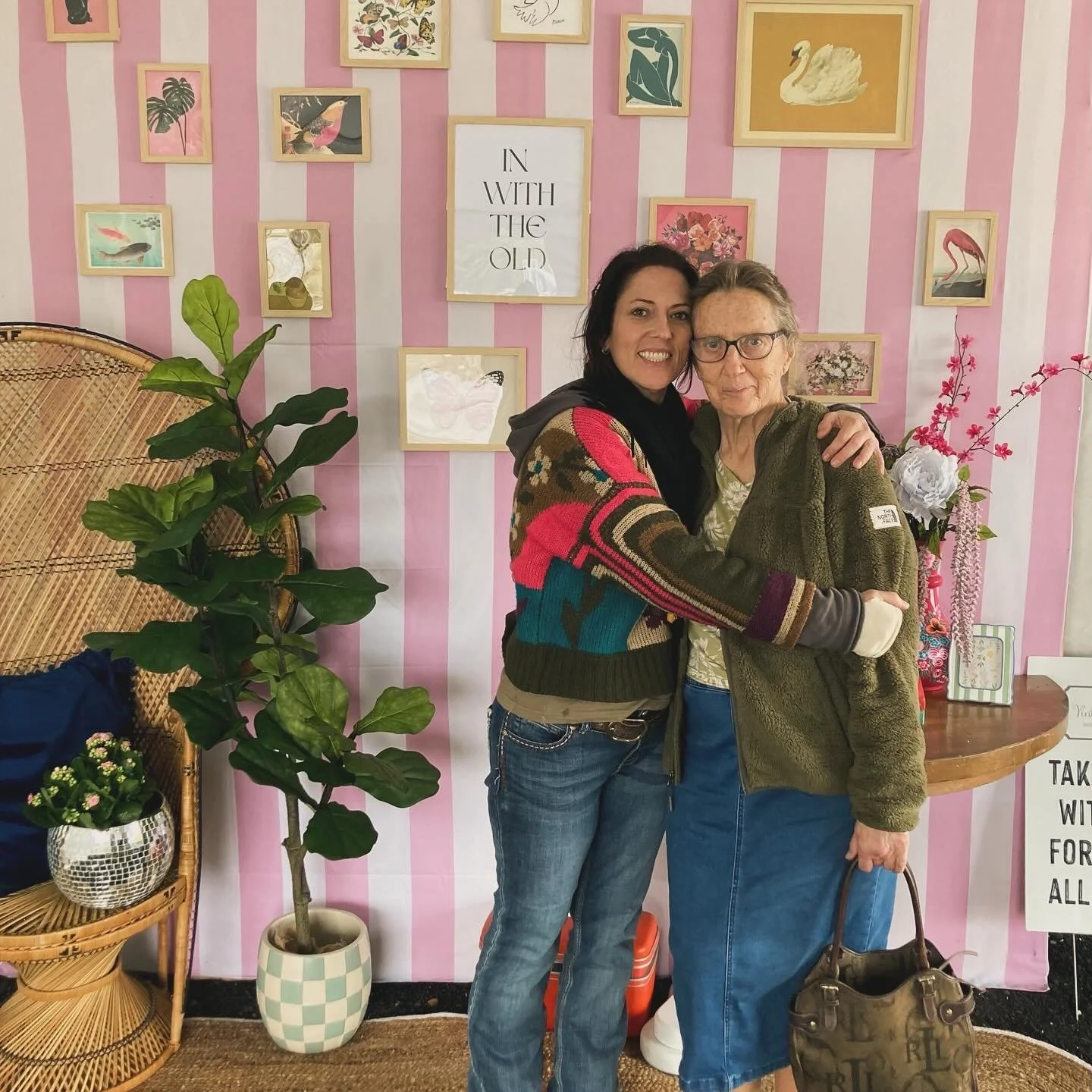Two women embracing and smiling at the camera in front of a pink and white striped wall decorated with framed pictures of animals and nature. One woman has dark hair and is wearing a colorful sweater, and the other has gray hair and glasses, wearing a green jacket and carrying a handbag.