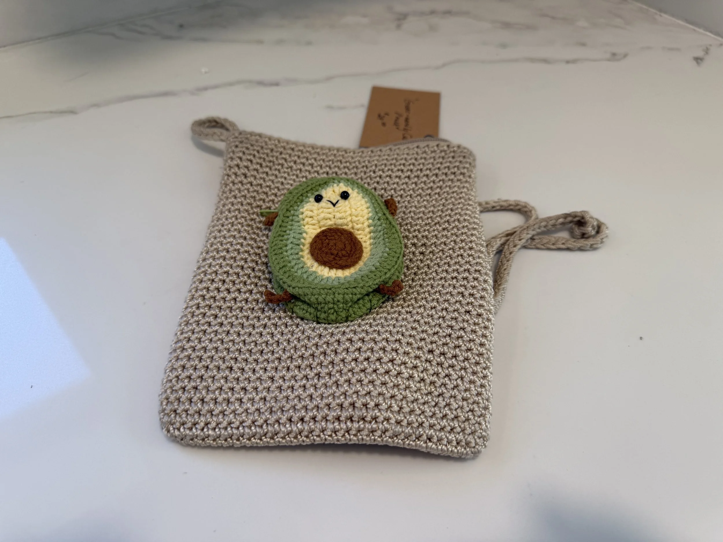 Knitted beige bag with a cute avocado face embroidery on it, placed on a white marble surface.