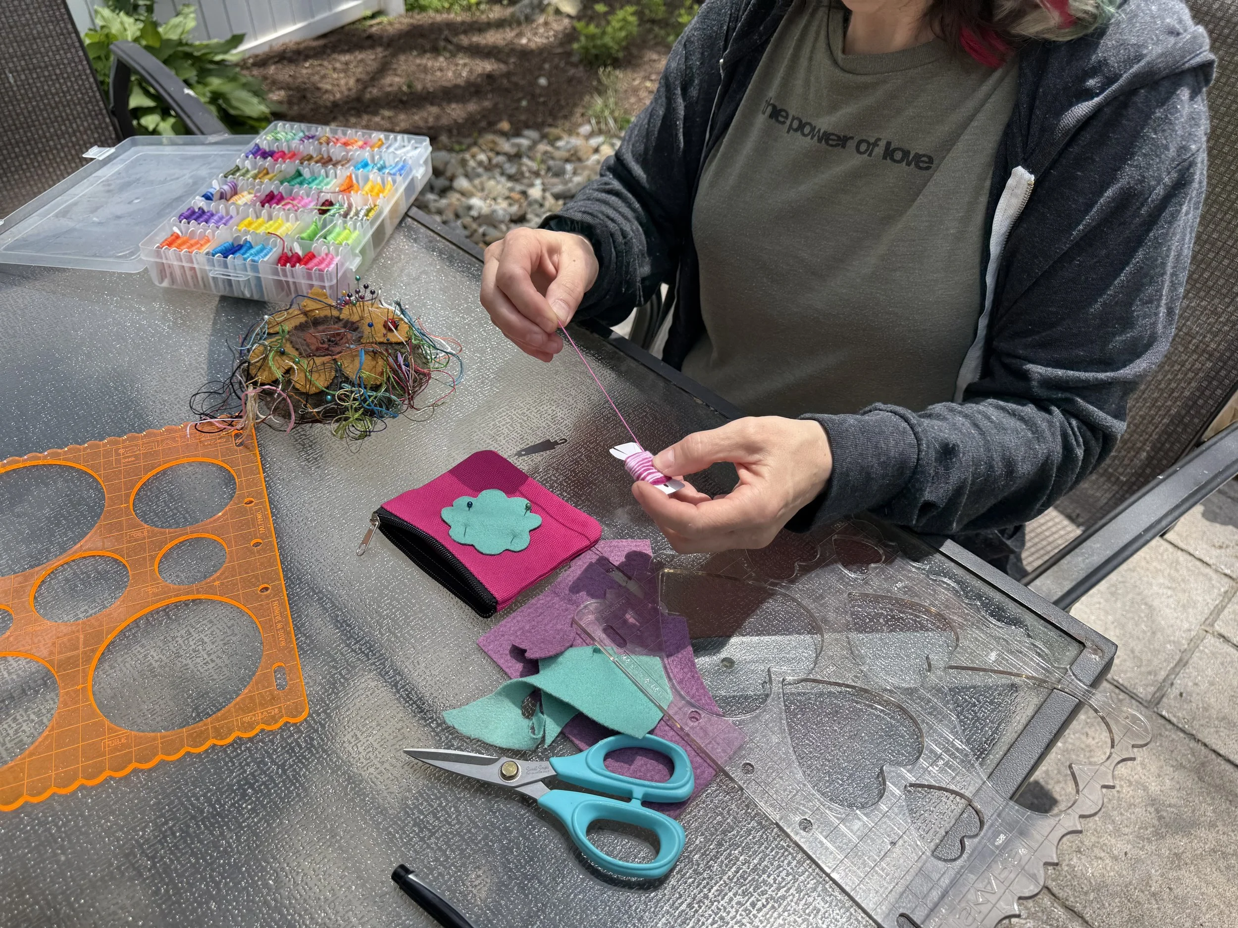 Person sewing buttons onto felt shapes at a table outdoors, with sewing supplies including thread, scissors, and a case of colorful push pins.