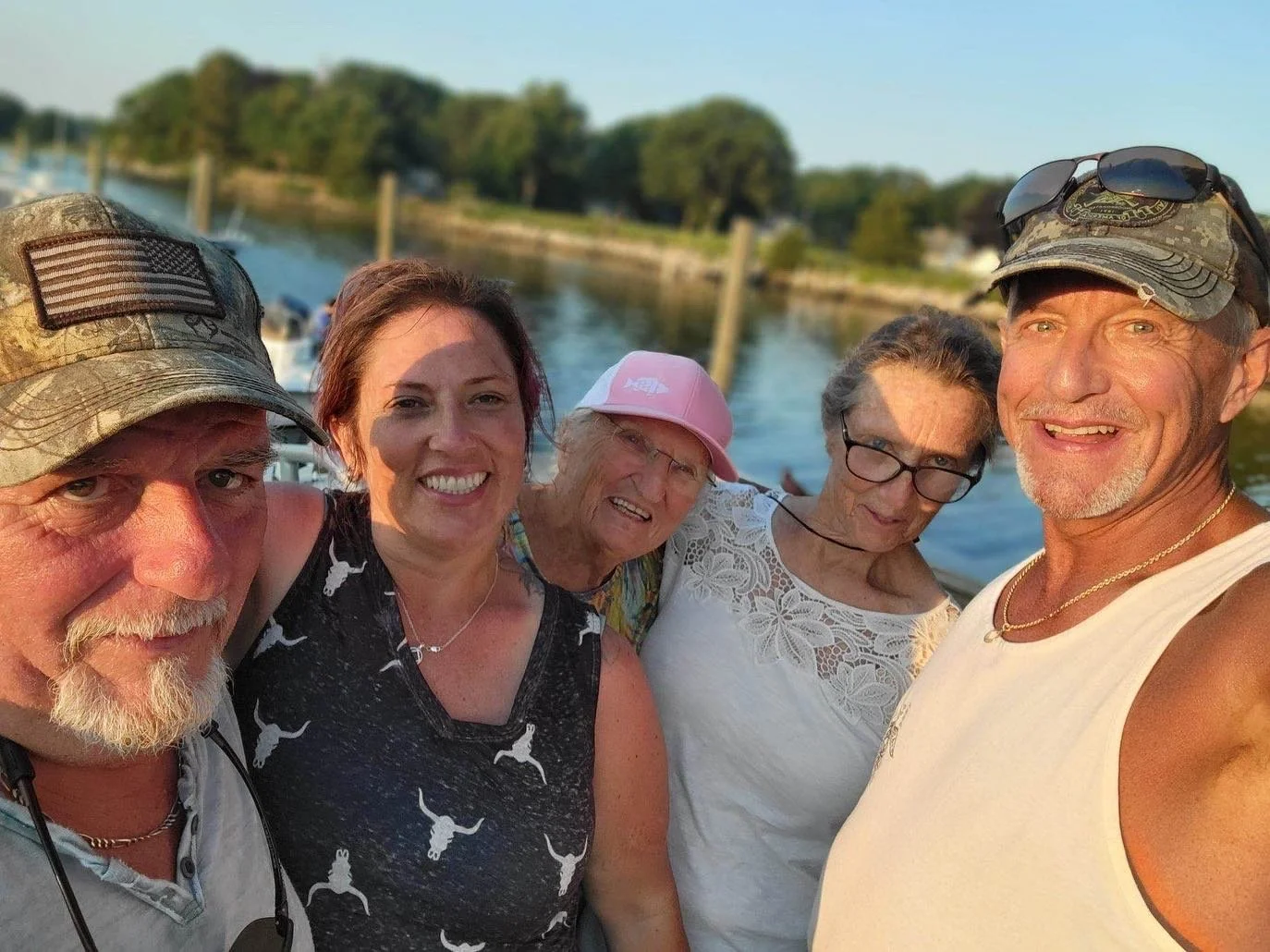 Group of five people taking a selfie outdoors near a body of water on a sunny day