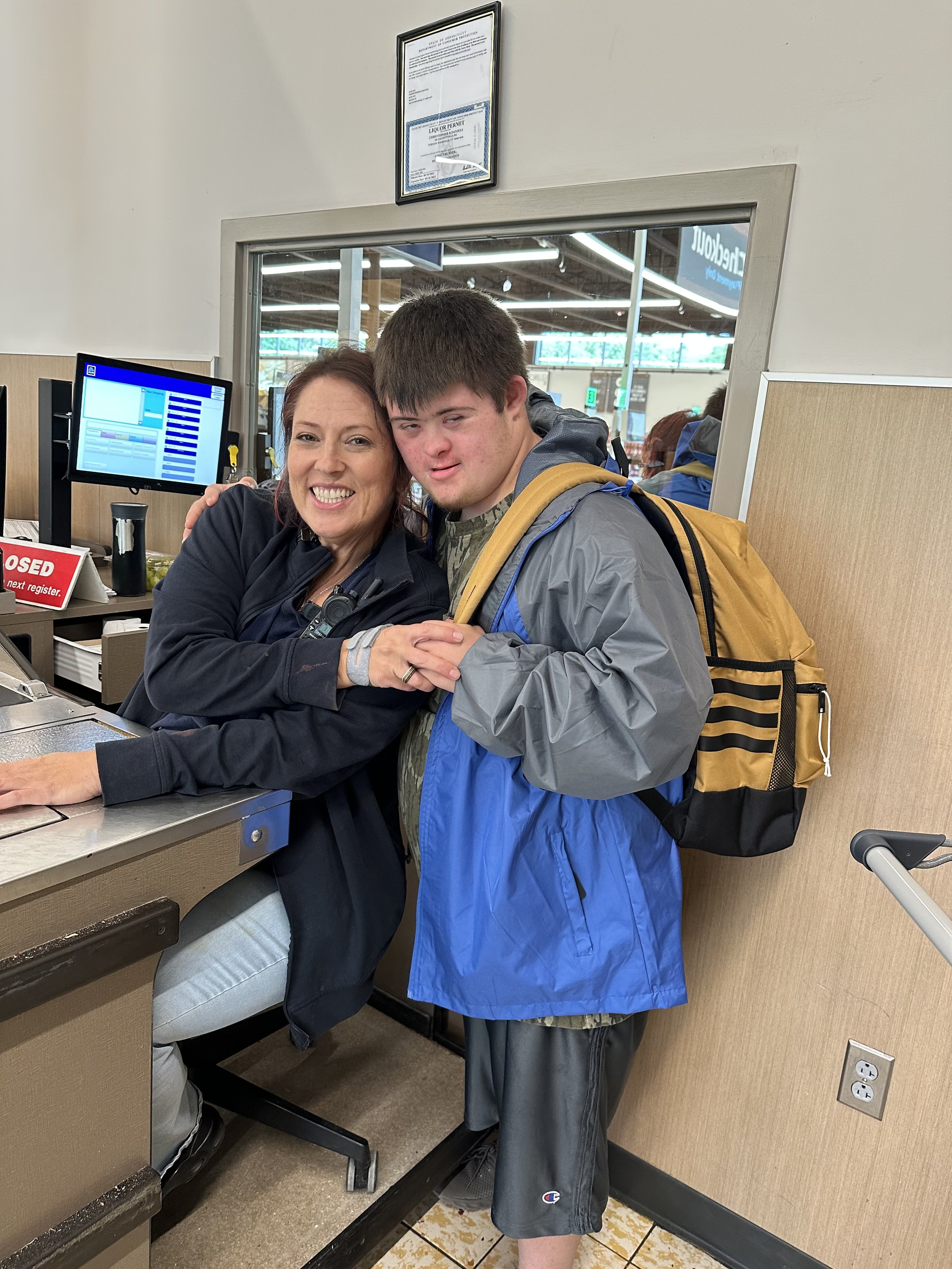 A woman and a teenage boy at a checkout counter in a store or restaurant. The woman is smiling and has her arm around the boy, who is wearing a backpack and looking at the camera. There is a monitor and some signage in the background.