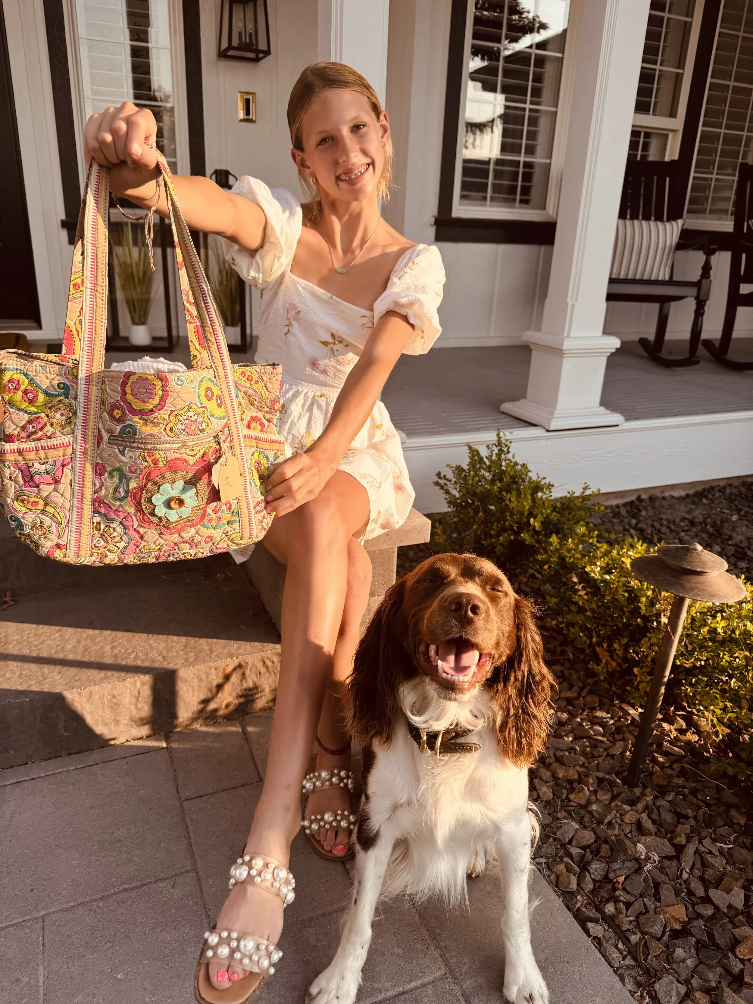 A young woman sitting on a step outside a house, holding a colorful bag and smiling at the camera. A happy brown and white dog stands in front of her. The house has white siding, black trim, and a porch with chairs, and the scene is lit by warm sunlight.
