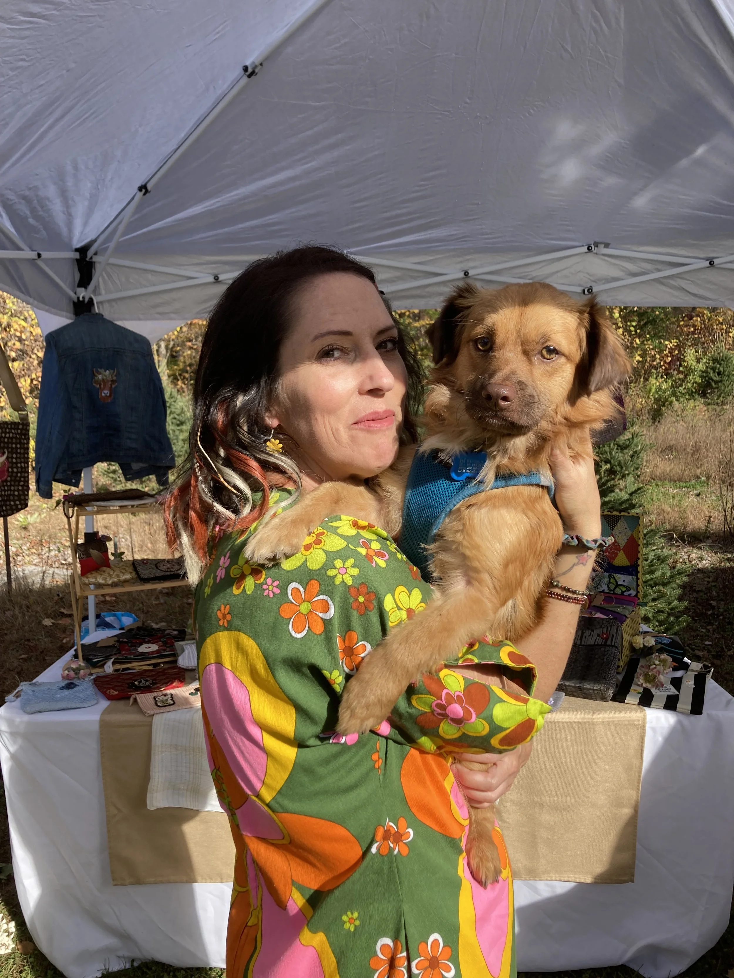 A woman with dark hair and colorful hair accents holding a brown dog with wearing a blue harness outdoors at a market or fair, with a white tent and tables with various items in the background.