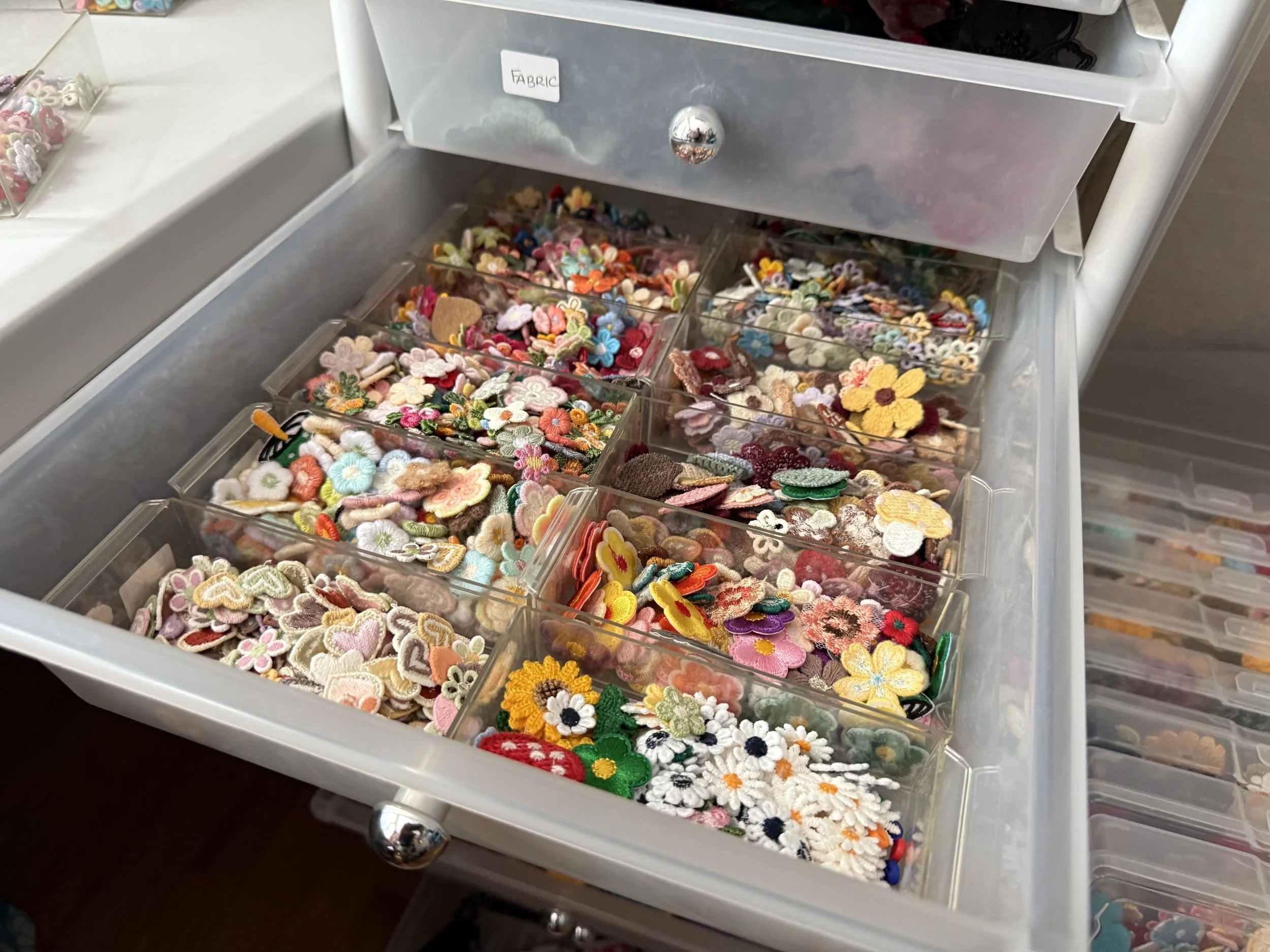 A white plastic drawer with multiple compartments filled with colorful embroidered and felt floral patches, some shaped like flowers and others with different designs.