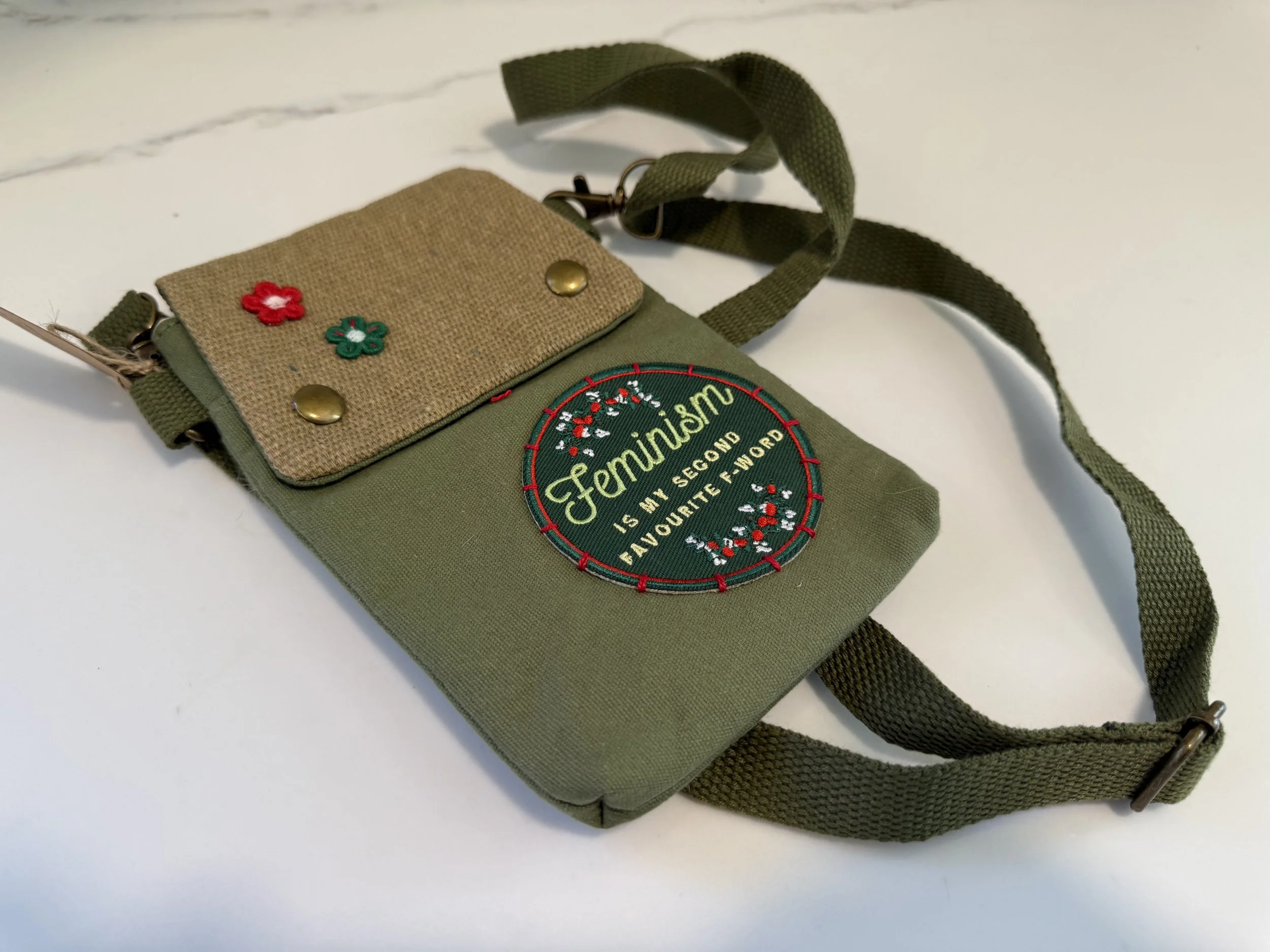 A small green fabric pouch with a beige flap, decorated with embroidered red and green flowers. It has a round embroidered patch that reads "Fermisn is my second favorite F-word." The pouch features metal snap closures and a green strap attached with metal hardware.