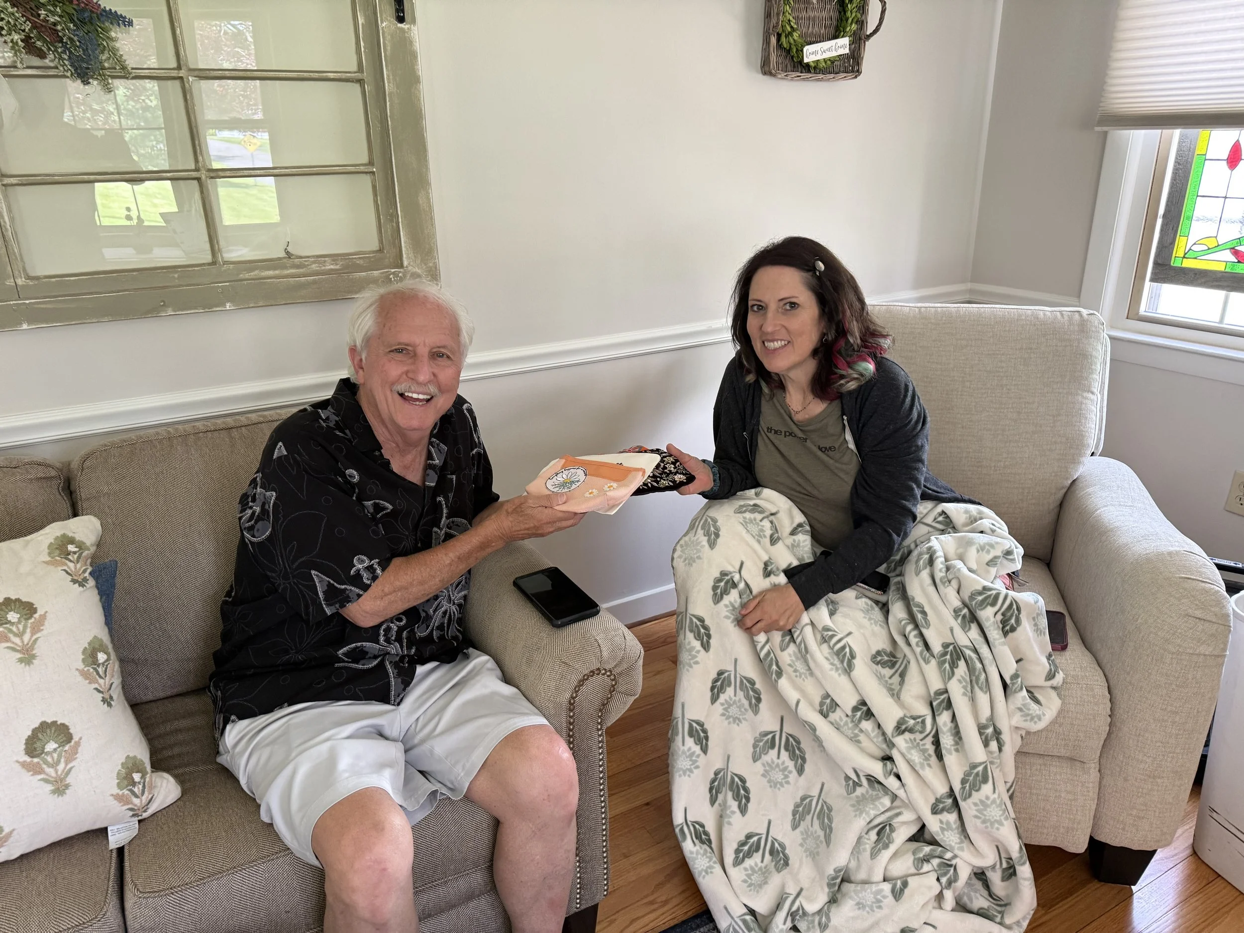 A smiling elderly man and a woman sitting on a beige couch in a bright room, exchanging items or presents. The man has white hair and is wearing a black patterned shirt and white shorts. The woman has dark hair with hints of pink, a gray T-shirt, and floral pajama pants. They are near a window with white blinds, and a stained glass window design is visible on the top right. The setting appears cozy and cheerful.