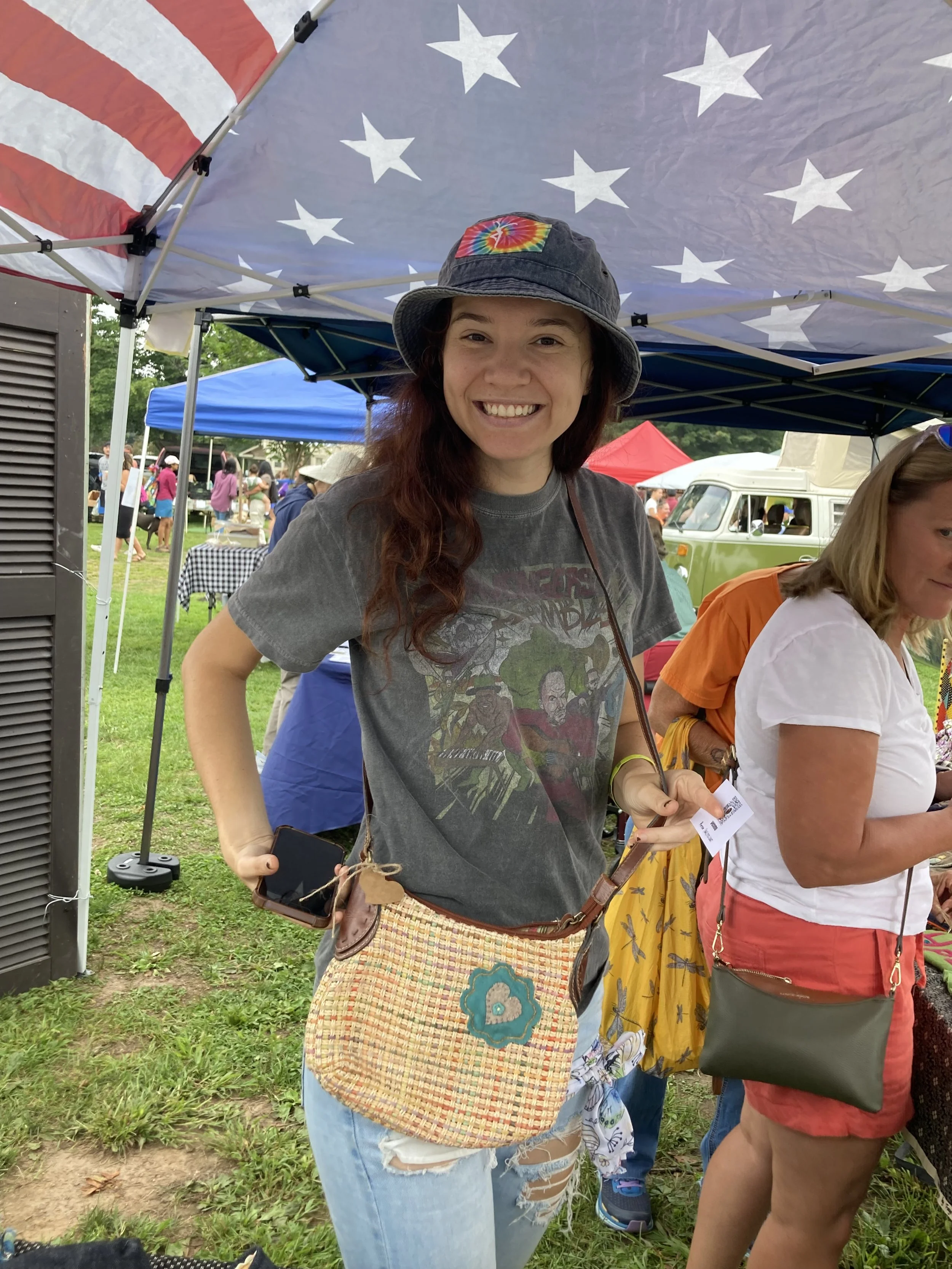 A smiling woman with long red hair wearing a tie-dye rainbow tie-dye bucket hat, a gray graphic T-shirt, and ripped jeans, holding a mobile phone and a ticket, standing under a tent with an American flag pattern. There are other people and tents in the background at an outdoor event.