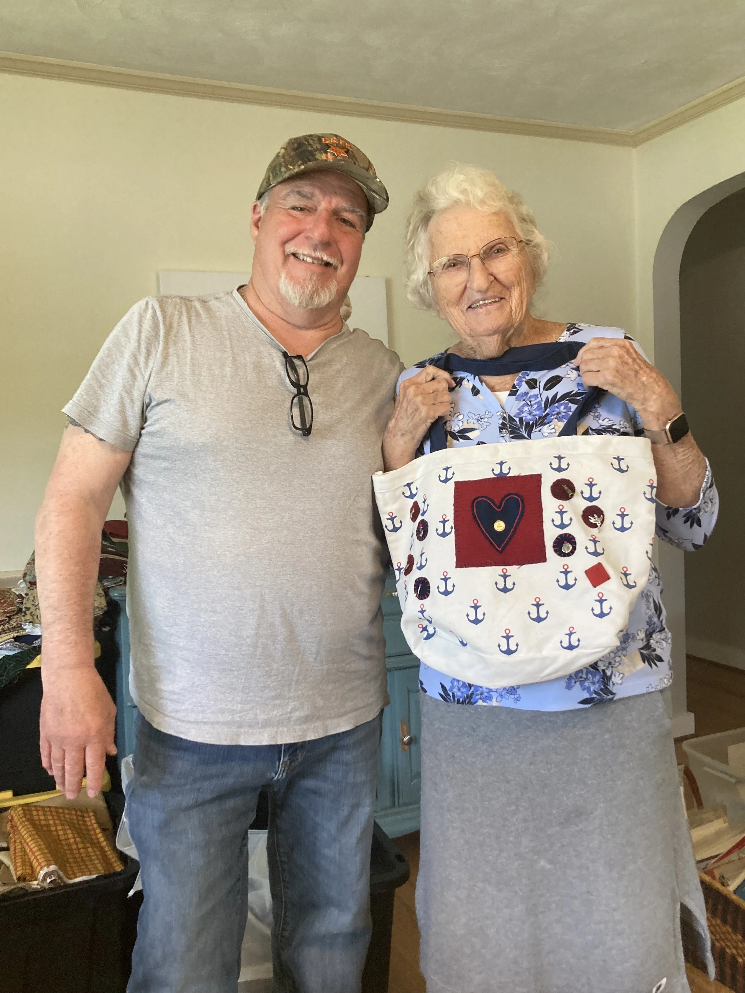 A man and an elderly woman smiling together indoors. The man is wearing a gray t-shirt, jeans, glasses hanging from his collar, and a baseball cap. The woman is wearing glasses, a blue floral top, and a gray skirt, holding a white tote bag with navy and red nautical designs.