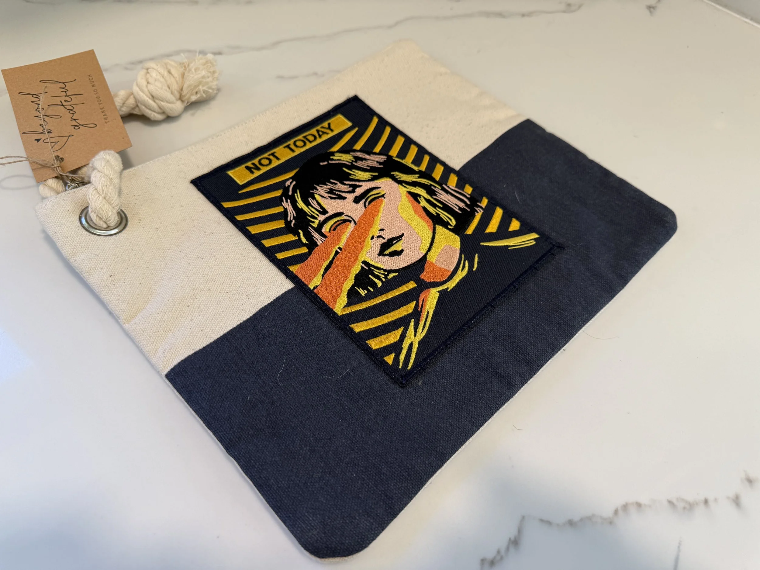 A fabric pouch with a colorful patch depicting a woman with hand over mouth and the text 'NOT TODAY' above her head, placed on a white surface.