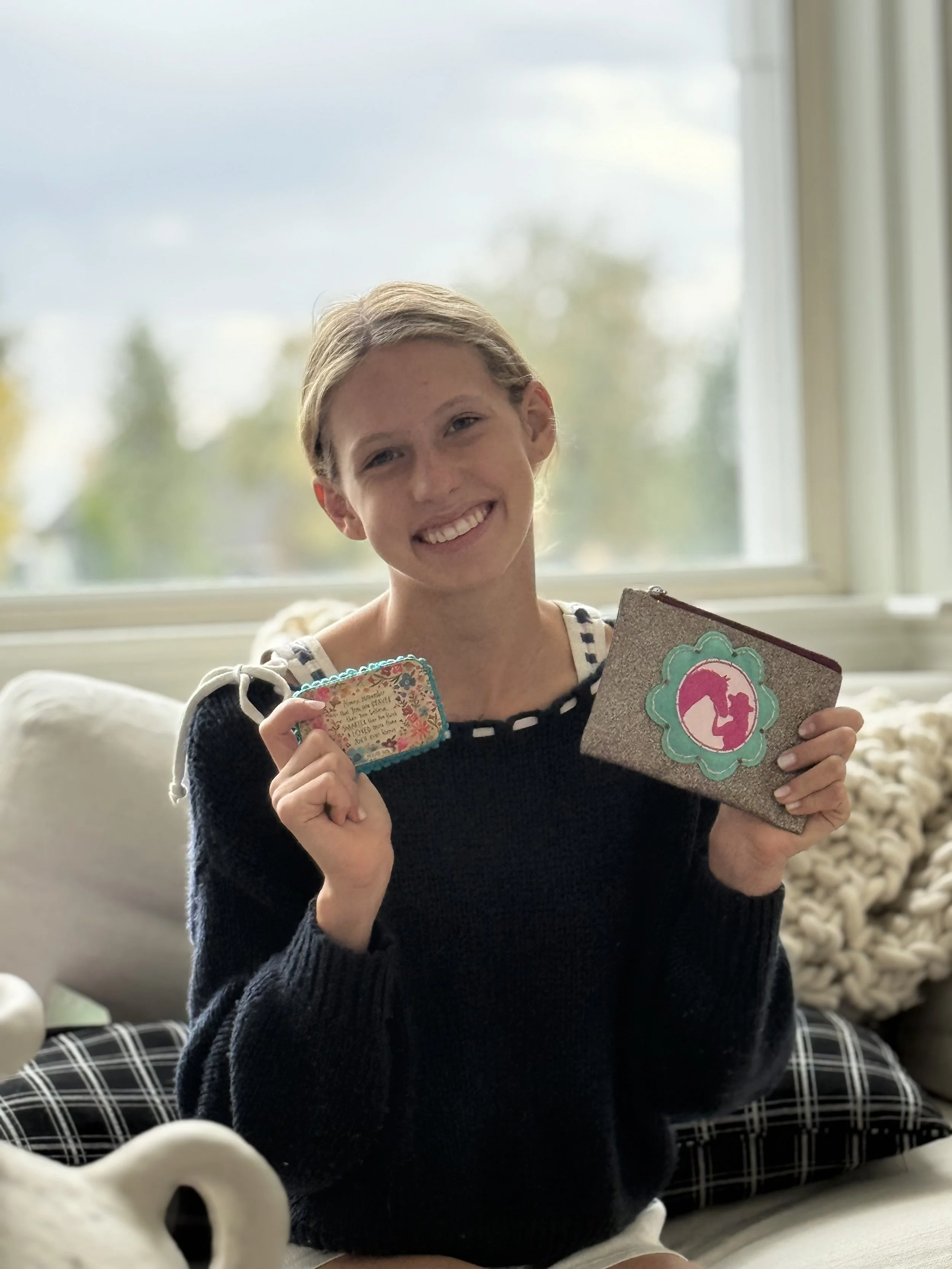 A young girl with blonde hair and a big smile holding up a decorated card and a smaller card inside her hands, sitting in front of a large window with a sunny outdoor view.