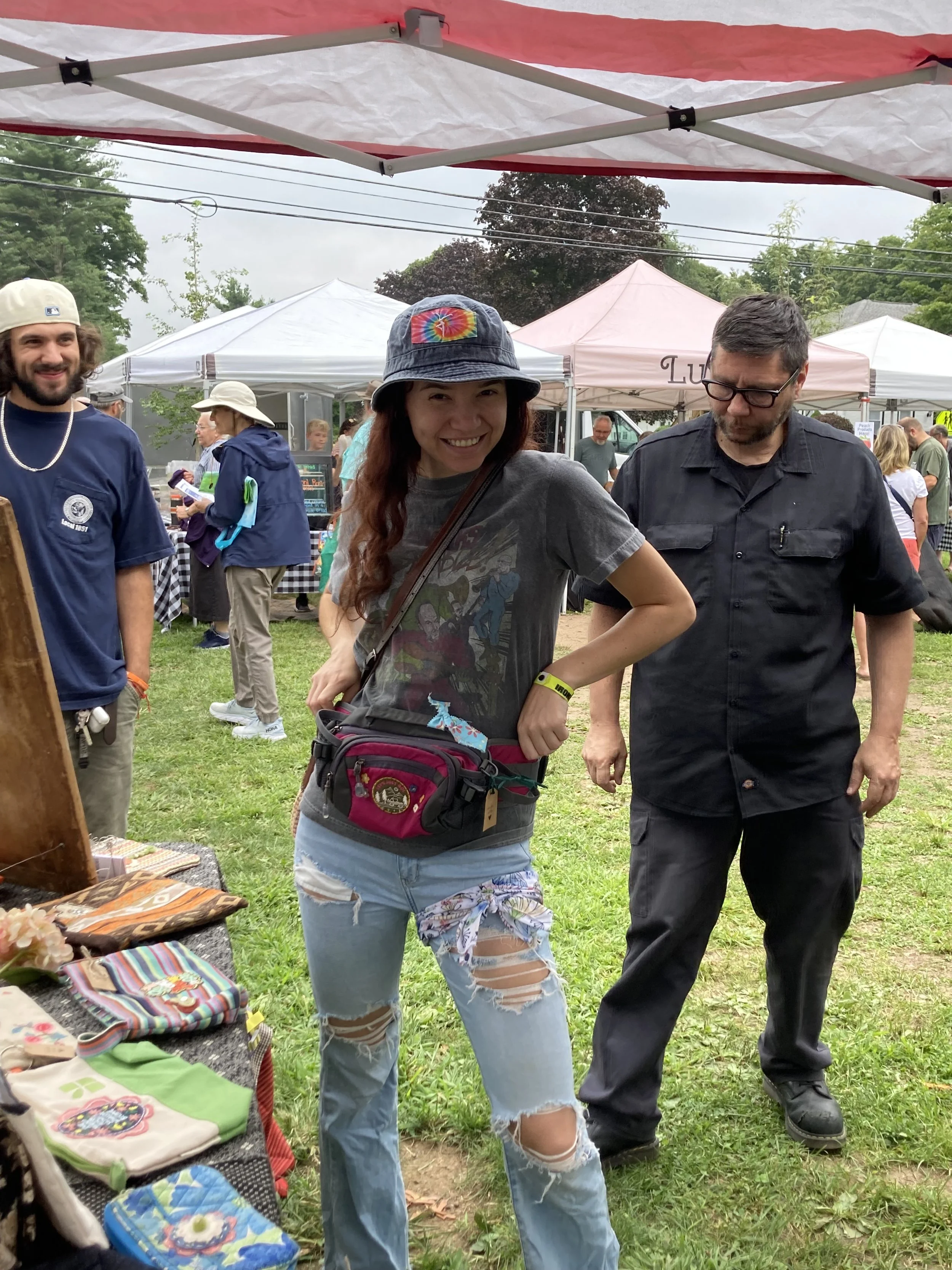 People attending an outdoor craft market or fair, tents, tables with handmade items, and greenery in the background, with overcast sky.