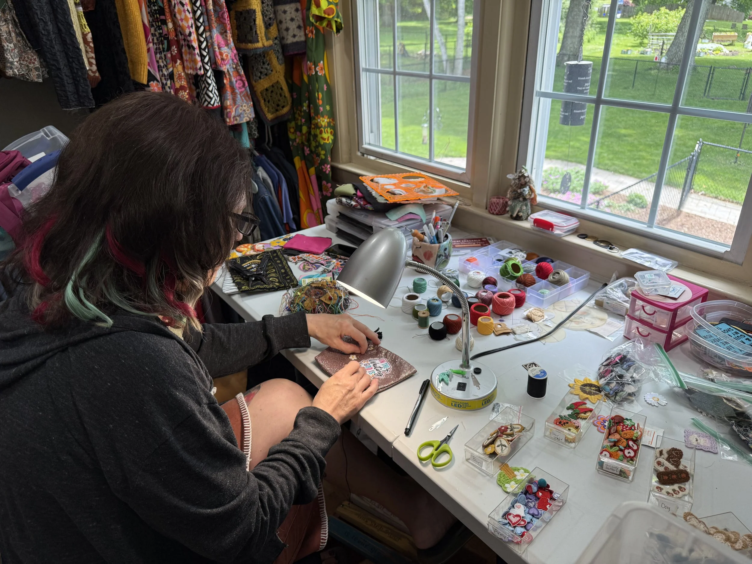 Woman working on crafting projects at a cluttered craft table with buttons, thread, scissors, and fabric, near a window overlooking a backyard.