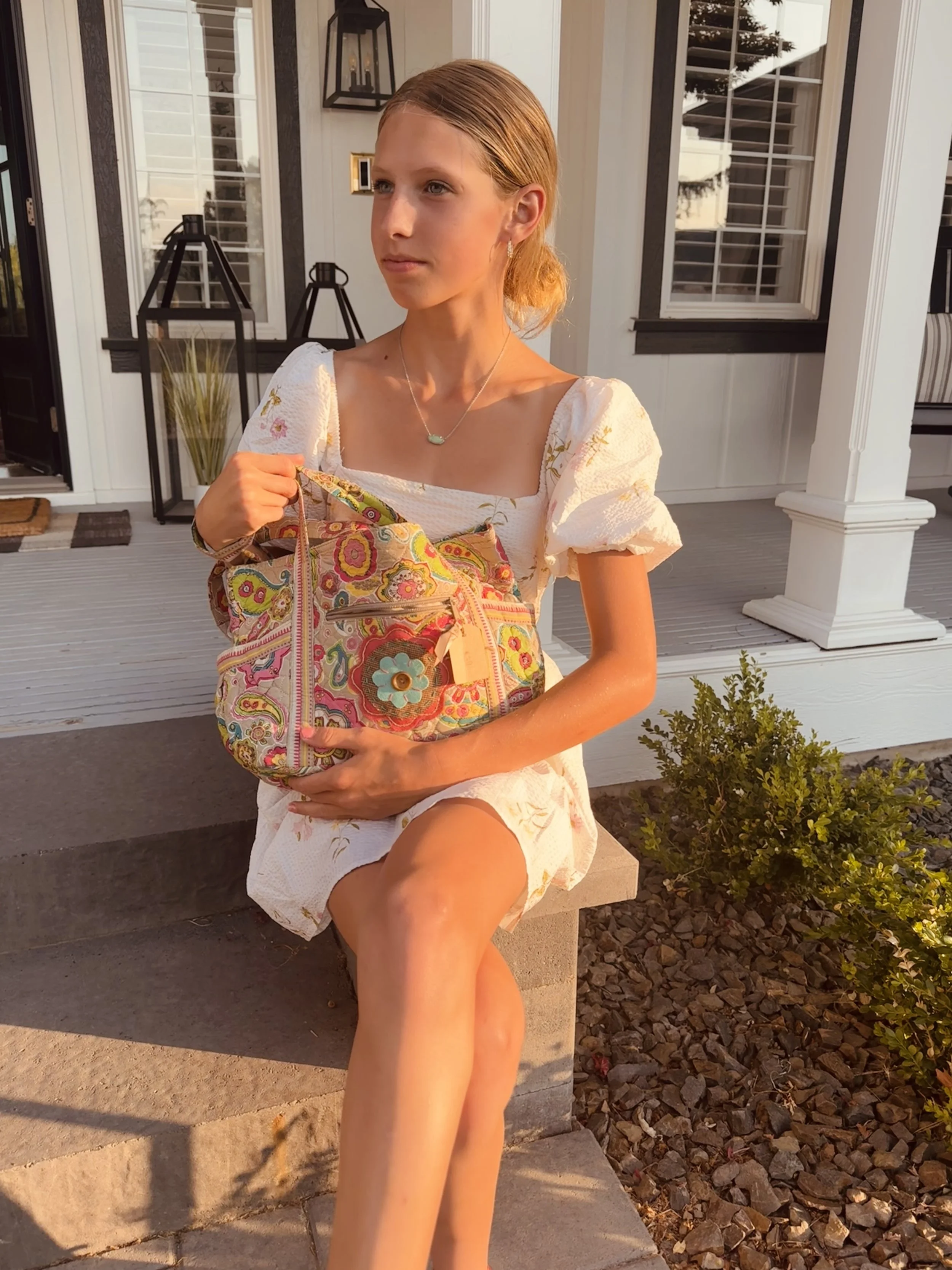 A young woman with red hair sits on a concrete step outside a house with white siding and black trim. She is wearing a white dress with sleeves and holding a colorful patterned bag. The sunlight is warm, casting a golden glow. There are potted plants and decorative lanterns on the porch.