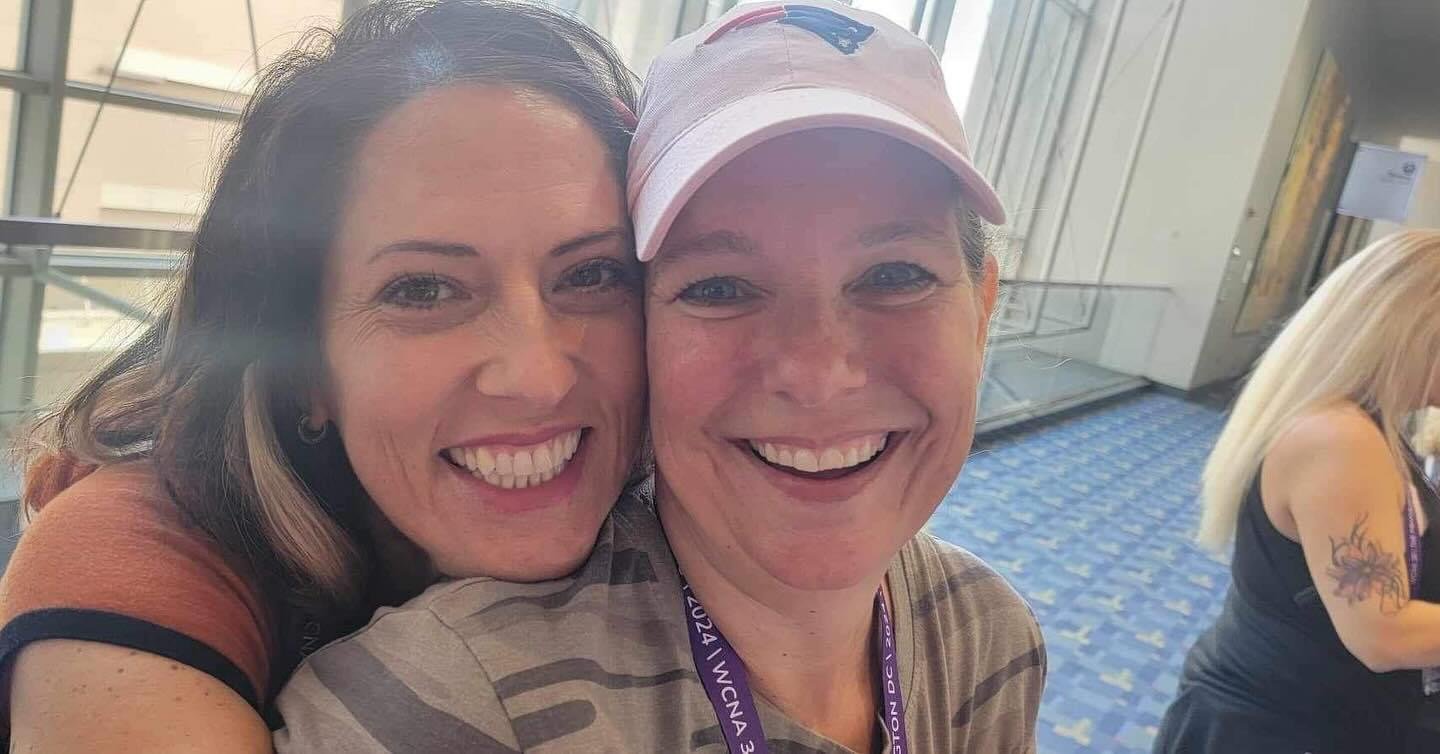 Two women smiling for a selfie at an indoor event, with one woman wearing a pink cap and the other with shoulder-length wavy hair.
