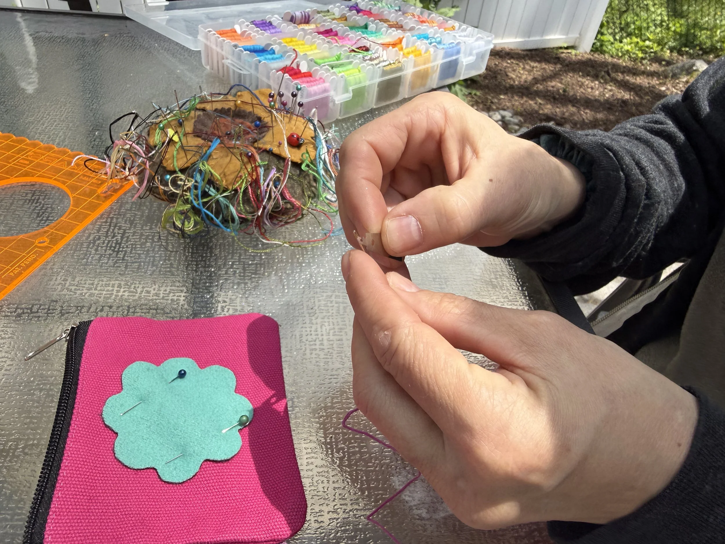 Person sewing decorative pins onto felt fabric outdoors, with a plastic organizer of colorful sewing pins in the background.