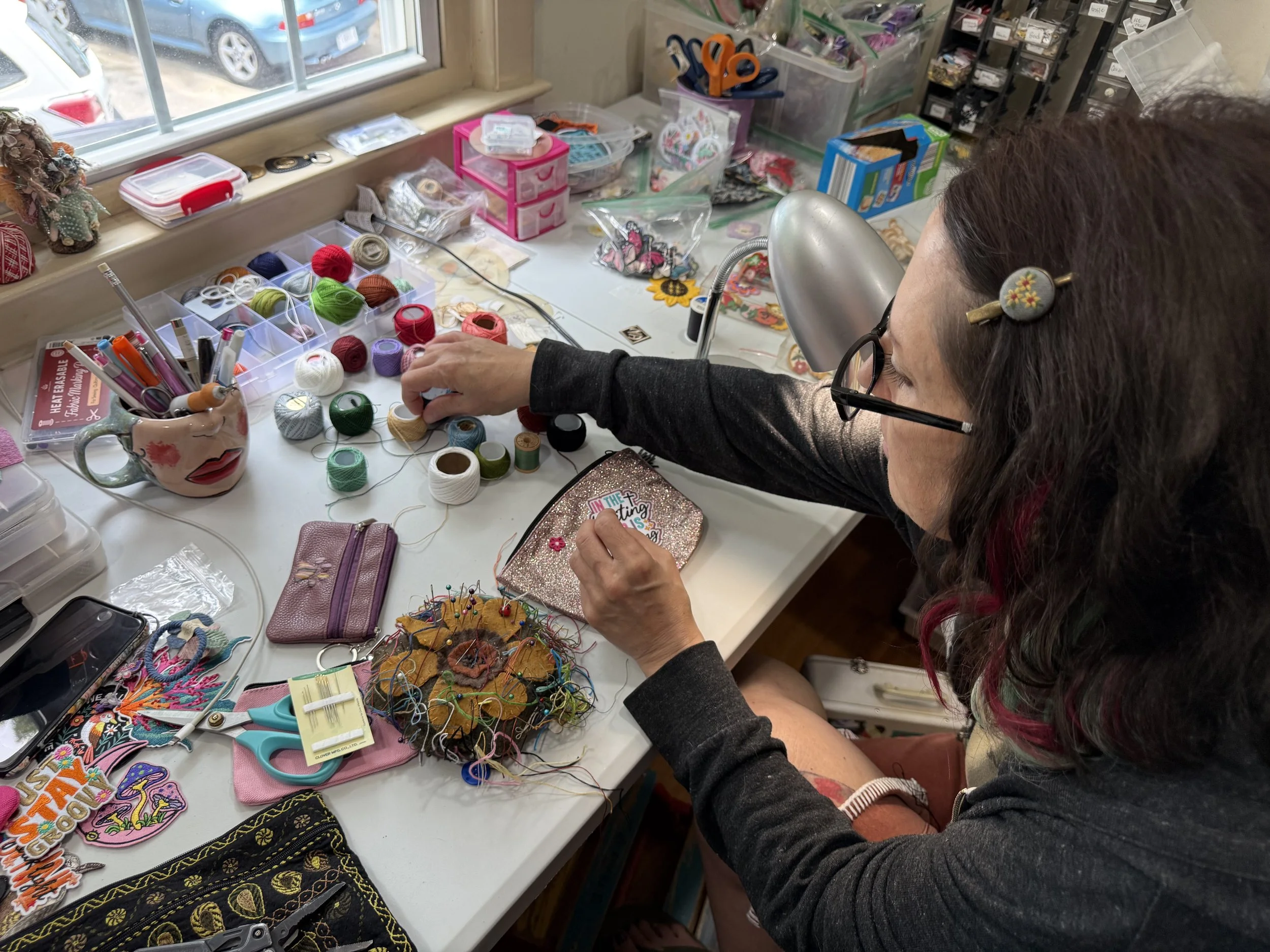 A woman with glasses and a floral hair clip is sitting at a cluttered craft table working with various sewing and embroidery supplies. The table has colorful threads, patches, scissors, and sewing tools, with additional supplies stored in plastic containers nearby. Outside the window, parked cars are visible.
