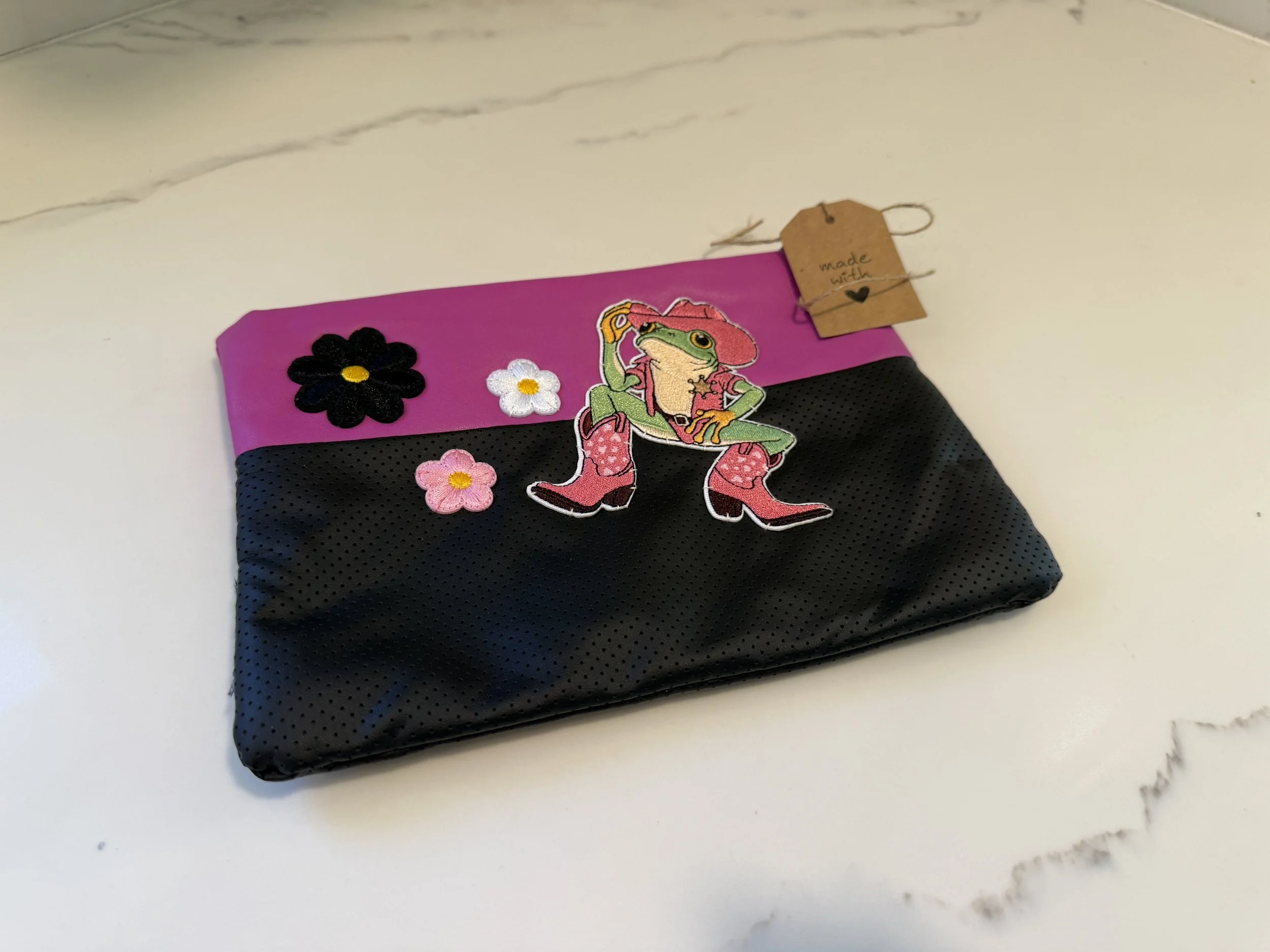 A pink and black fabric pouch decorated with embroidered patches including a dinosaur wearing pink boots, and flower patches in black, white, and pink. A brown paper tag attached with string reads 'made with' and a small heart. The pouch is placed on a white marble surface.