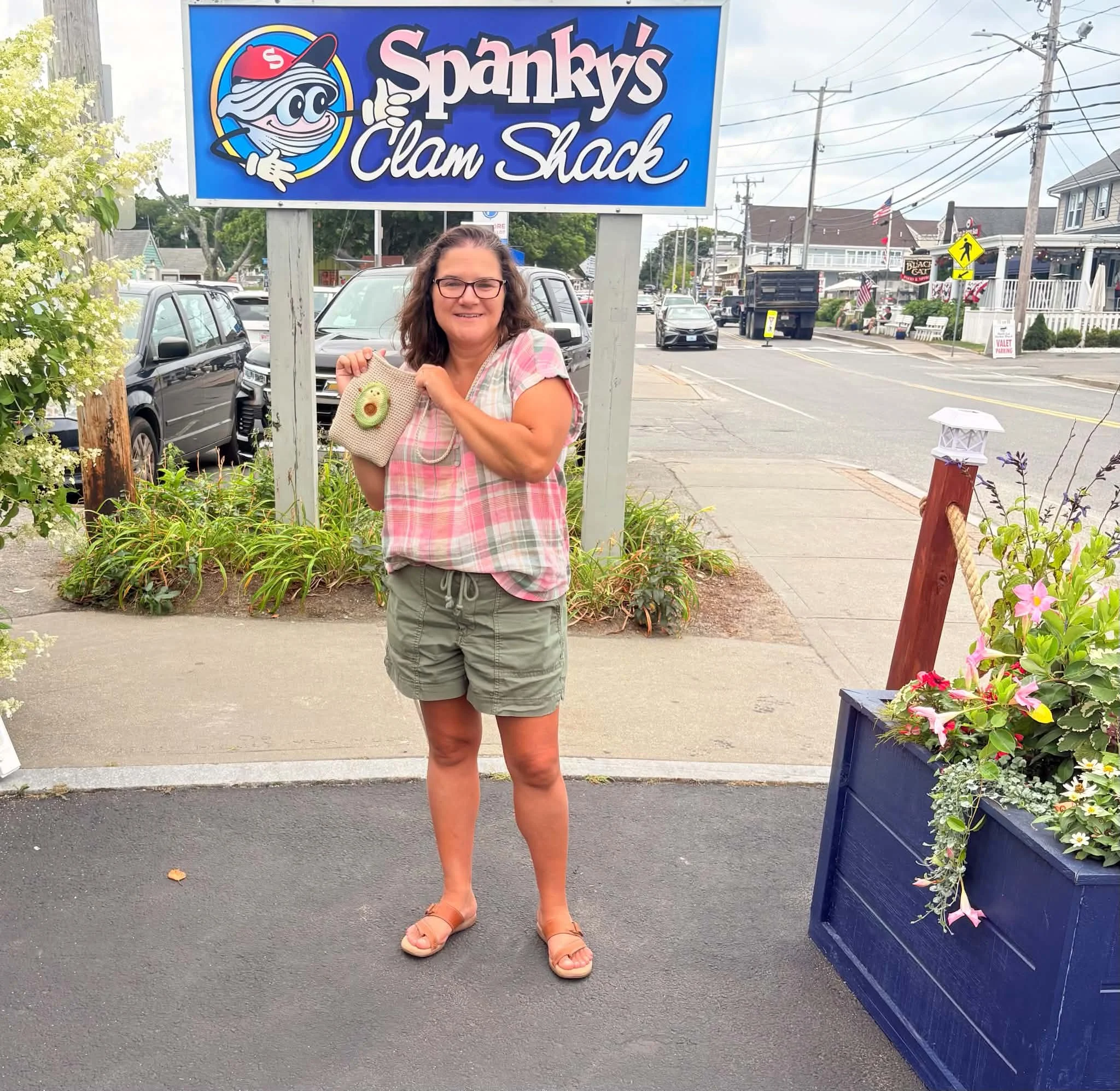 Woman standing outside at the sidewalk, smiling, holding a beige handbag with a green and brown decorative piece, in front of a sign for Sparky's Clam Shack. The background shows parked cars, buildings, and utility poles.