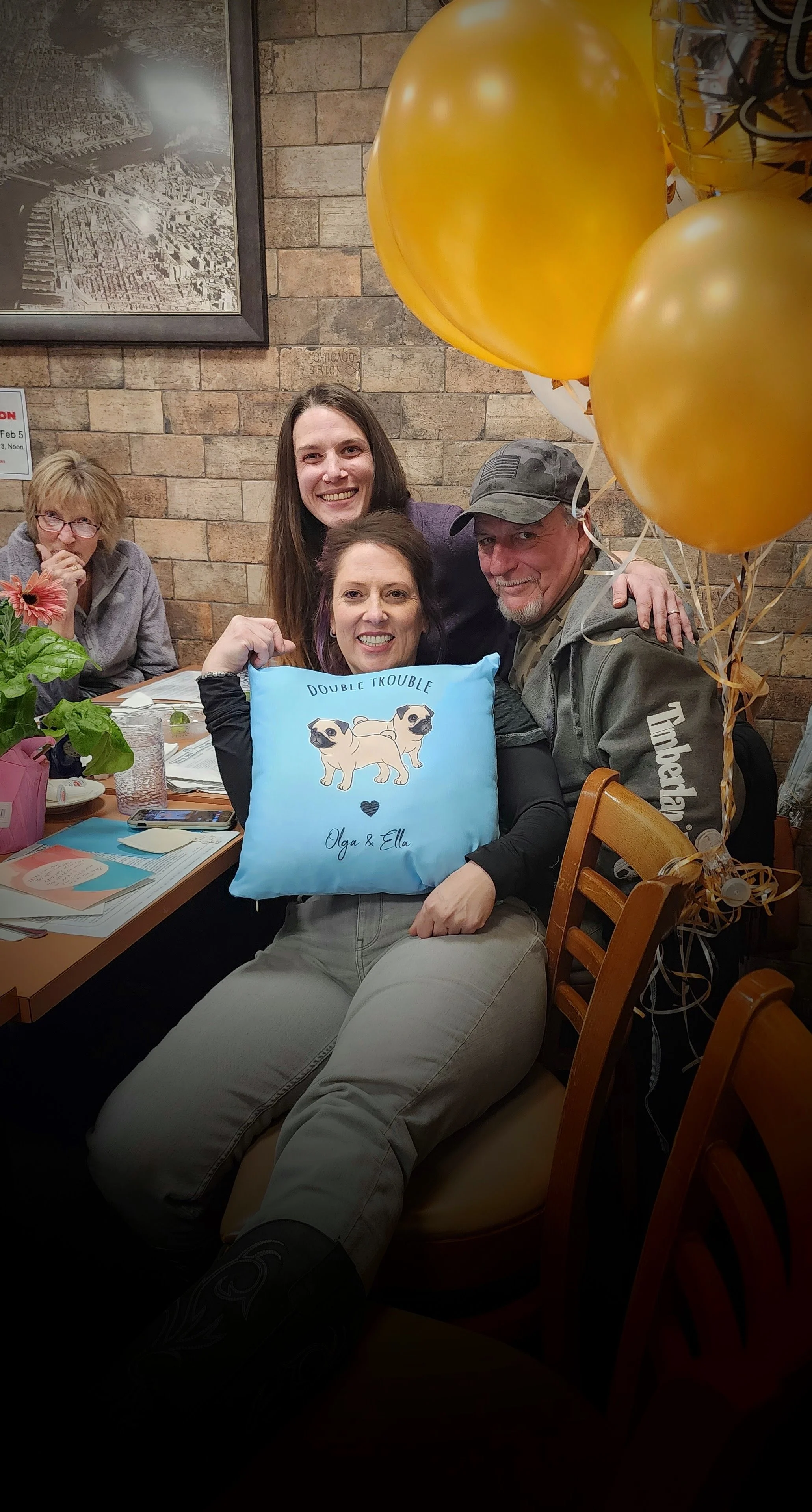 Group of four people sitting at a restaurant table, with two women in the foreground holding a blue cushion with pug dogs and the words 'Double Trouble' and 'Olga & Ella.' The woman on the left is wearing black and the woman on the right is wearing a gray baseball cap. The background features a brick wall, framed black and white cityscape photo, and gold balloons.
