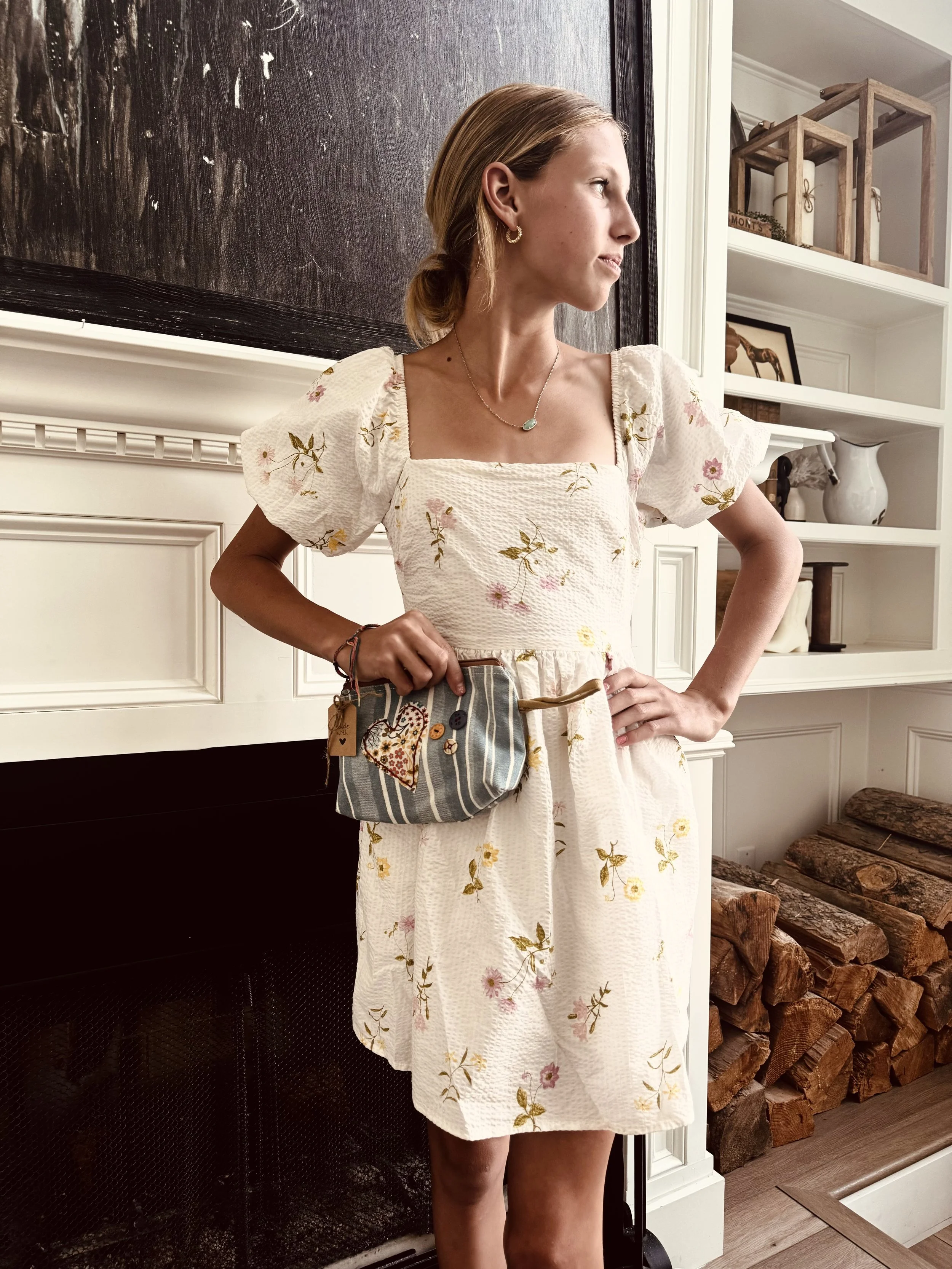 A woman with blond hair tied back, wearing a white floral dress with puffed sleeves, jewelry including earrings and a necklace, is standing indoors near a fireplace, looking to her right with her hands on her hips.