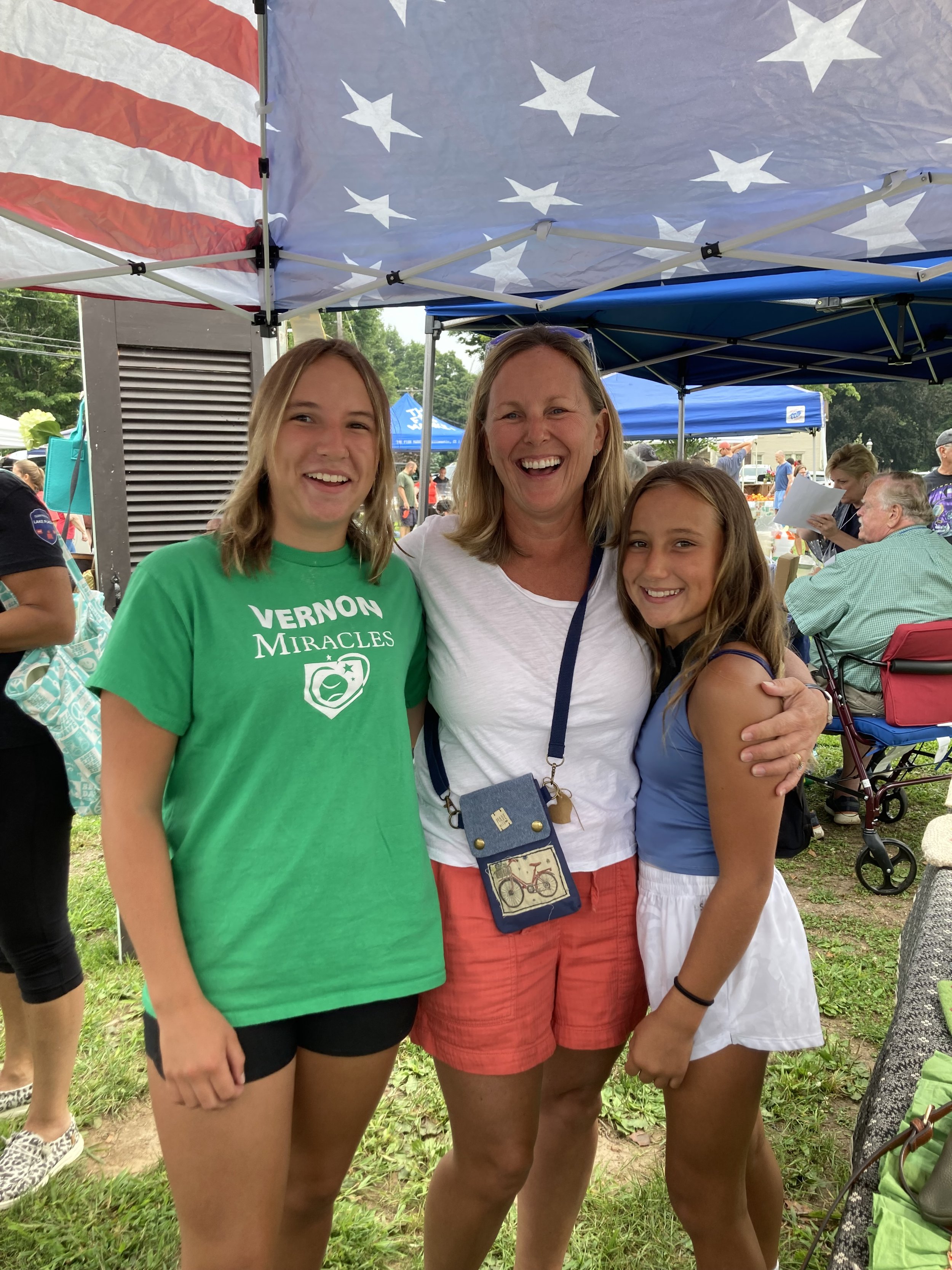 Three women smiling and posing outdoors under a canopy with an American flag pattern, surrounded by people and tents at what appears to be a community event.