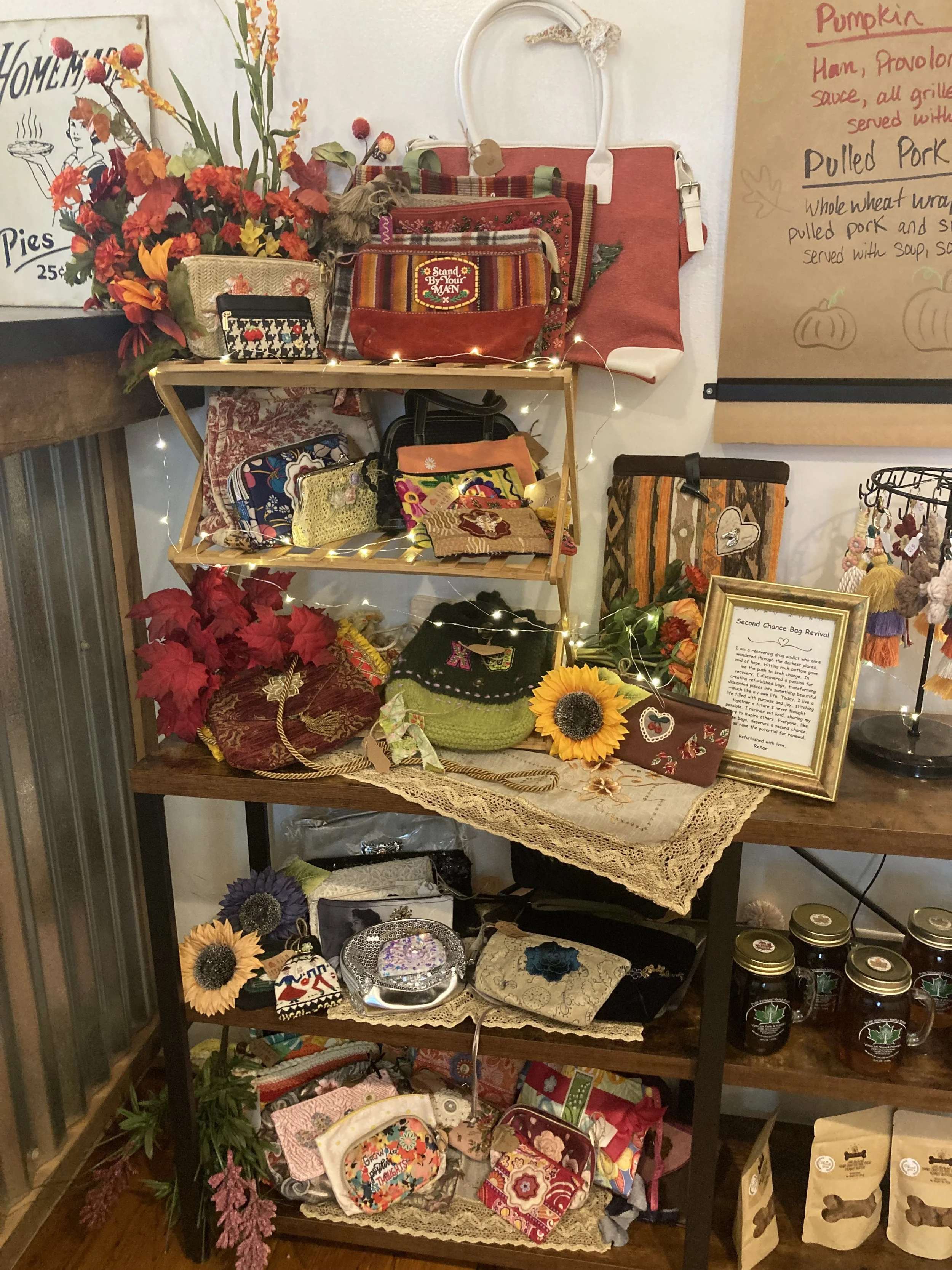 A display shelf with handmade and decorative items including handbags, sunflower decorations, and small gift boxes. There are intricate fabrics, a framed note, and various craft projects arranged on the shelves adorned with string lights and artificial flowers.